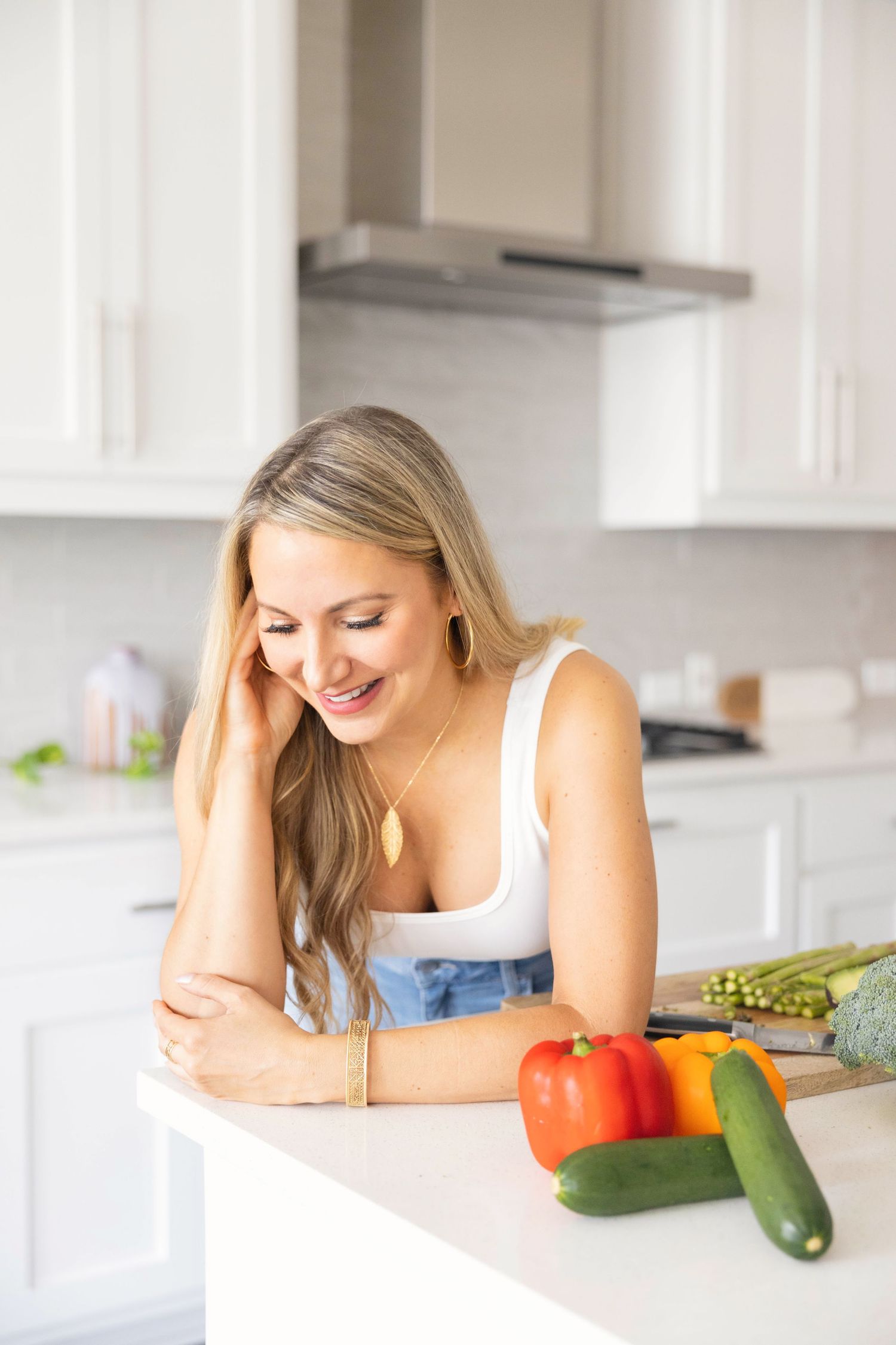 Blonde woman in white tank top smiling while leaning on white kitchen counter with fresh vegetables.
