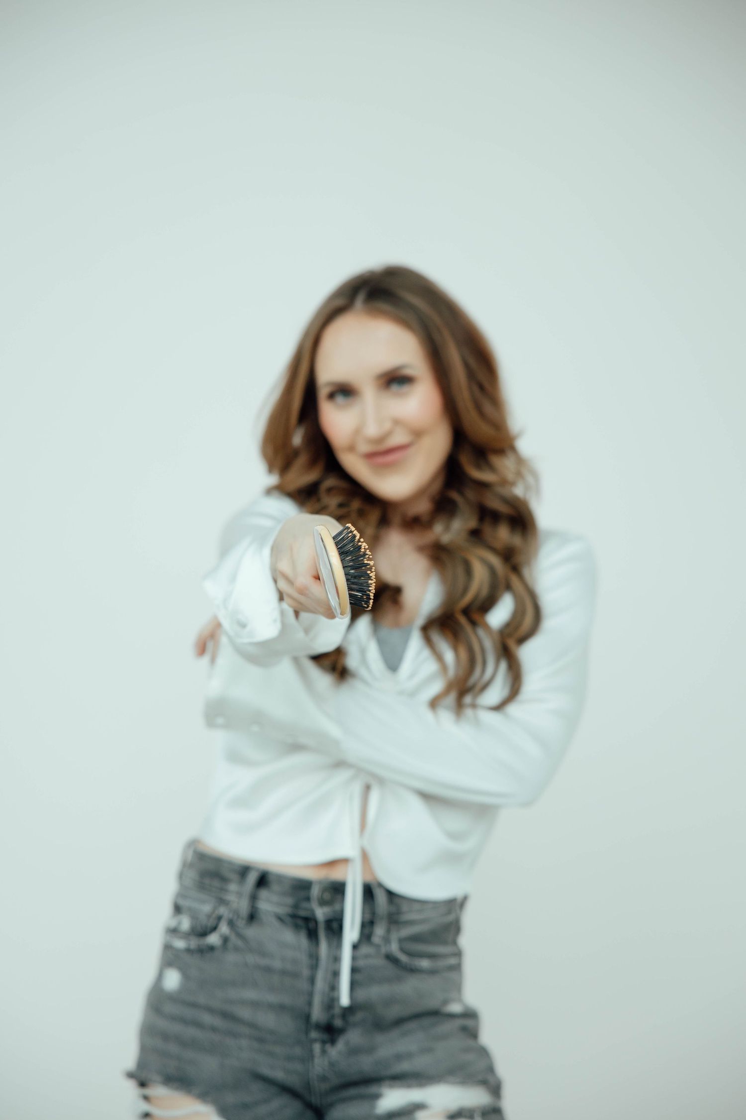 Brunette woman in white top and distressed jeans pointing at camera against light background, bracelet in focus.