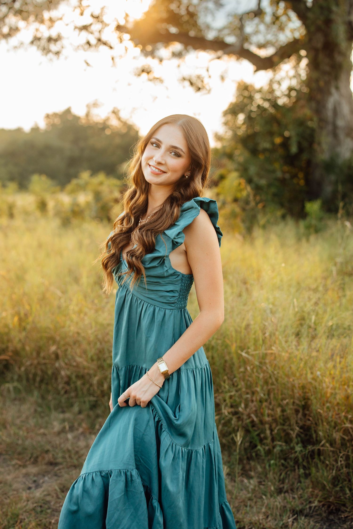 A young woman in a teal ruffled maxi dress smiles while standing in a sunlit meadow.