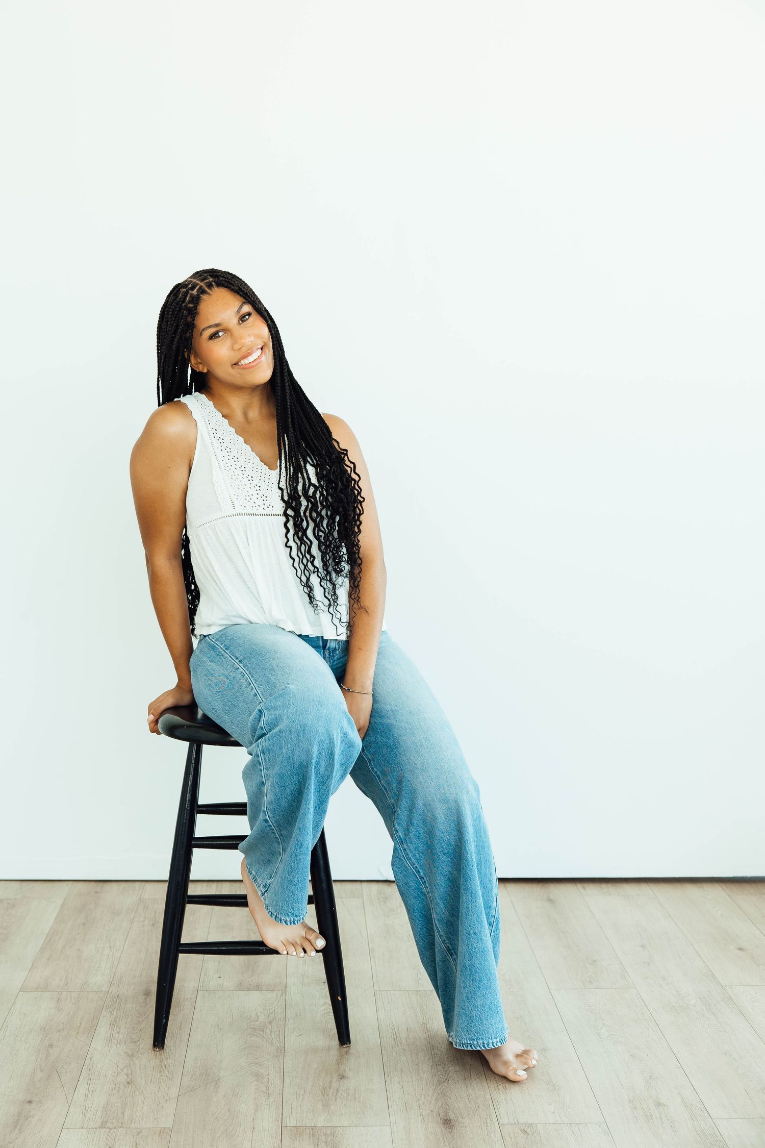 Young Black woman with long braids in white top and wide-leg jeans sitting on a wooden stool.
