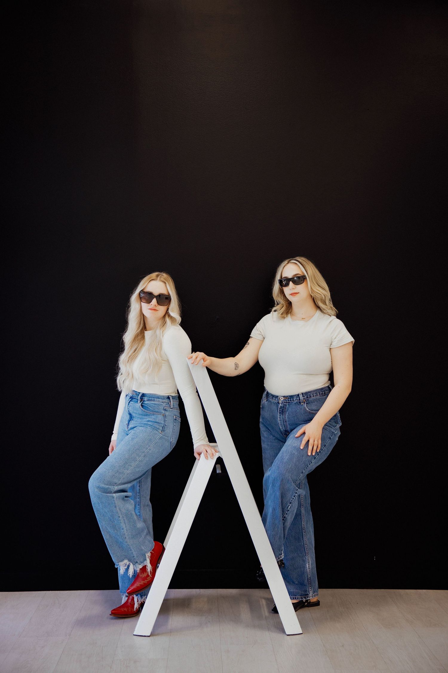 Two blonde women in white tees and jeans pose with white ladder against black studio wall.