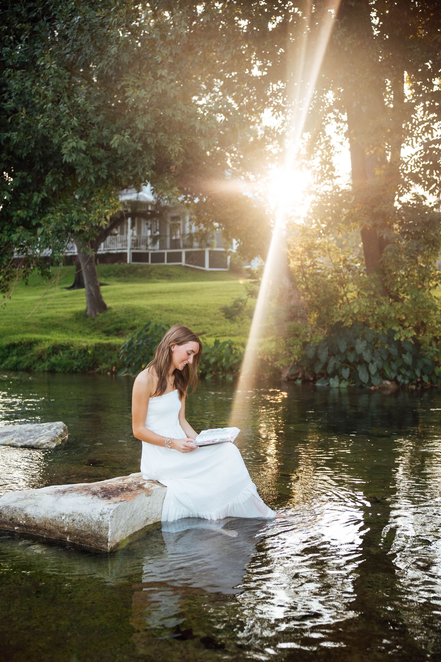 Young woman in white dress reading on rocks by a sunlit creek.