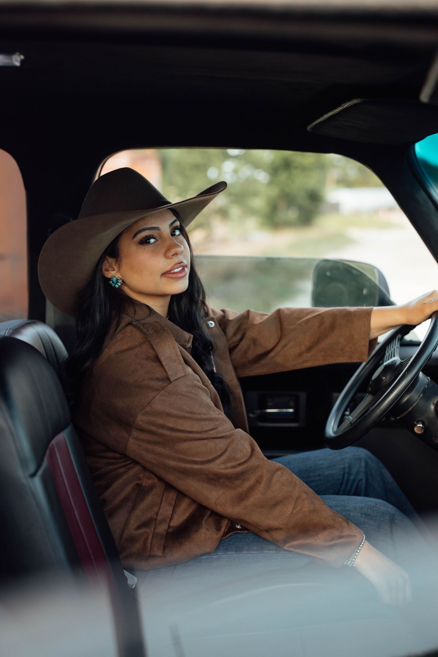 A woman in a brown suede jacket and cowboy hat sits in the driver's seat of a vintage truck.