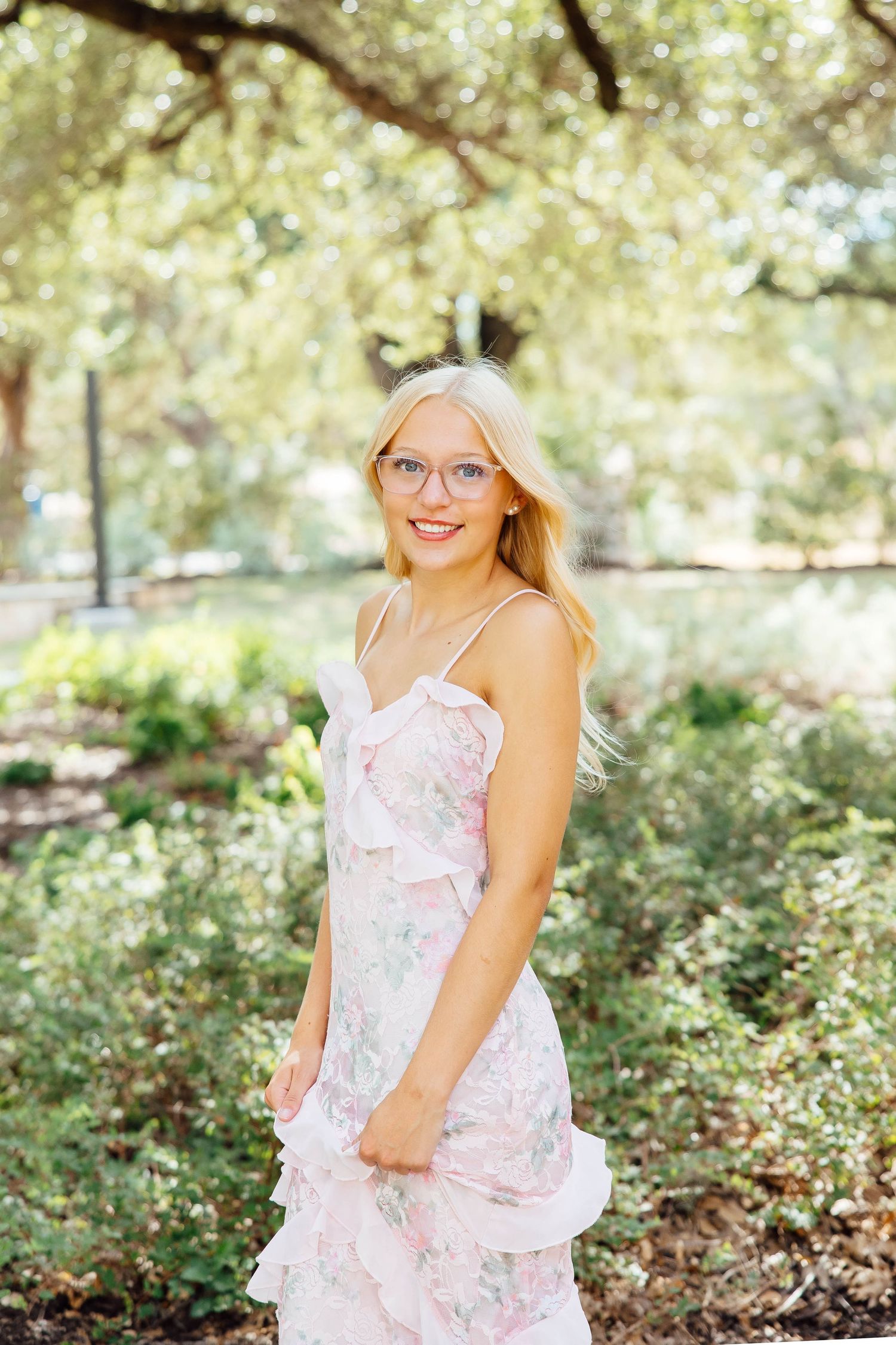 Smiling blonde woman in pink floral dress with glasses stands in lush green garden setting.