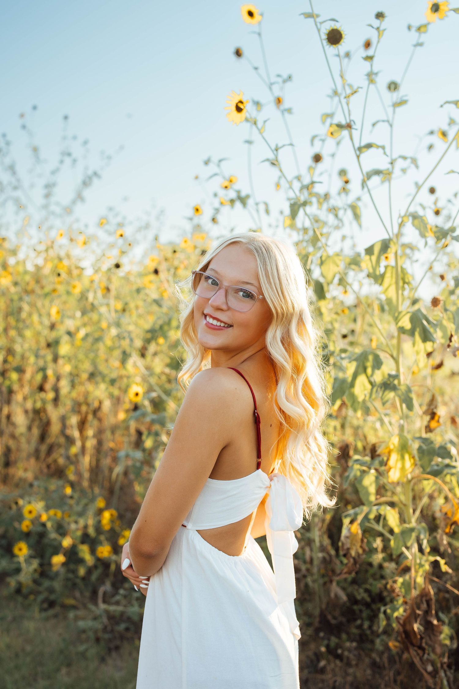Smiling blonde woman with glasses in white backless dress standing in sunflower field at sunset.