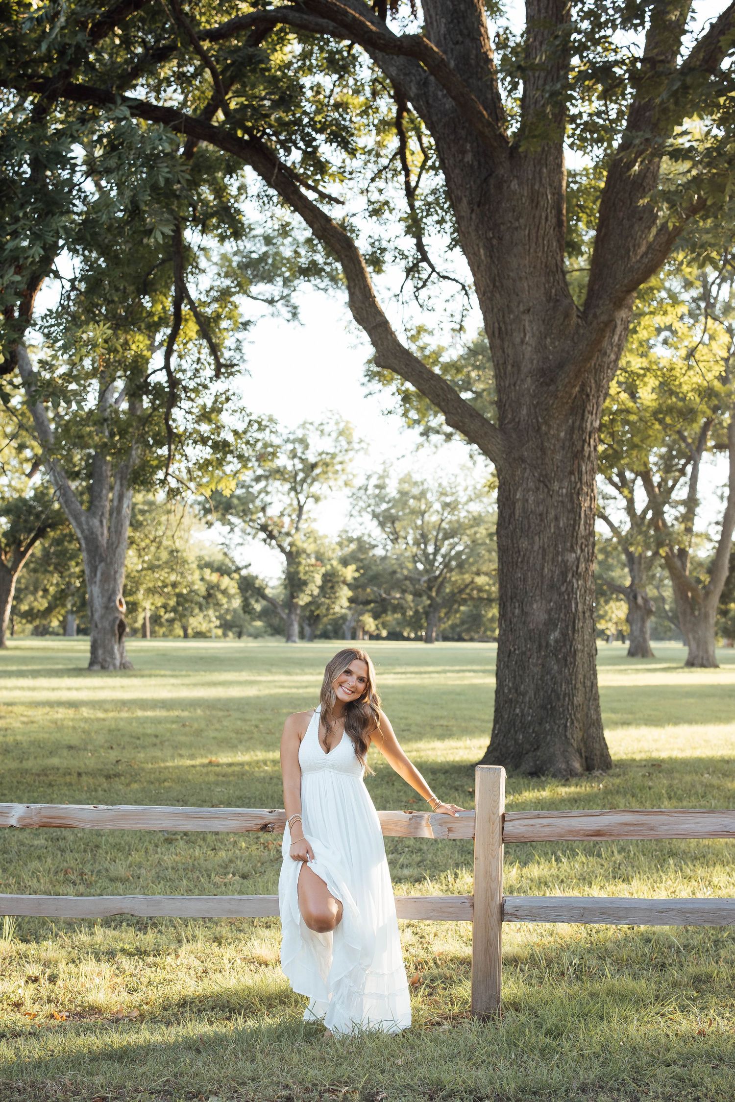 Woman in white maxi dress standing by wooden fence under large oak trees in sunny park.