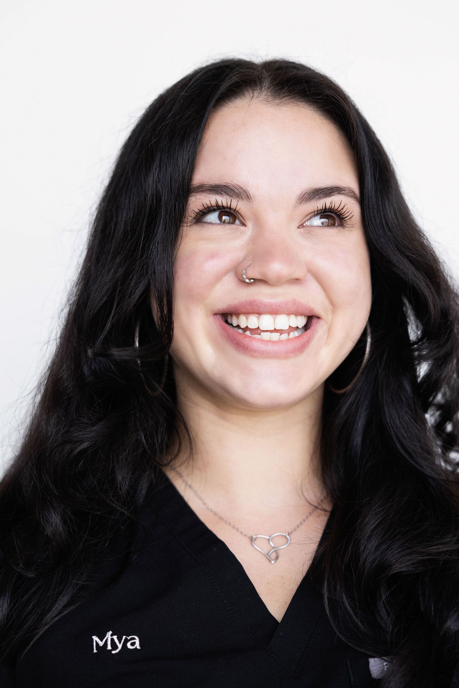 Young woman named Mya in black scrubs with dark wavy hair, nose ring, and hoop earrings smiling upward.