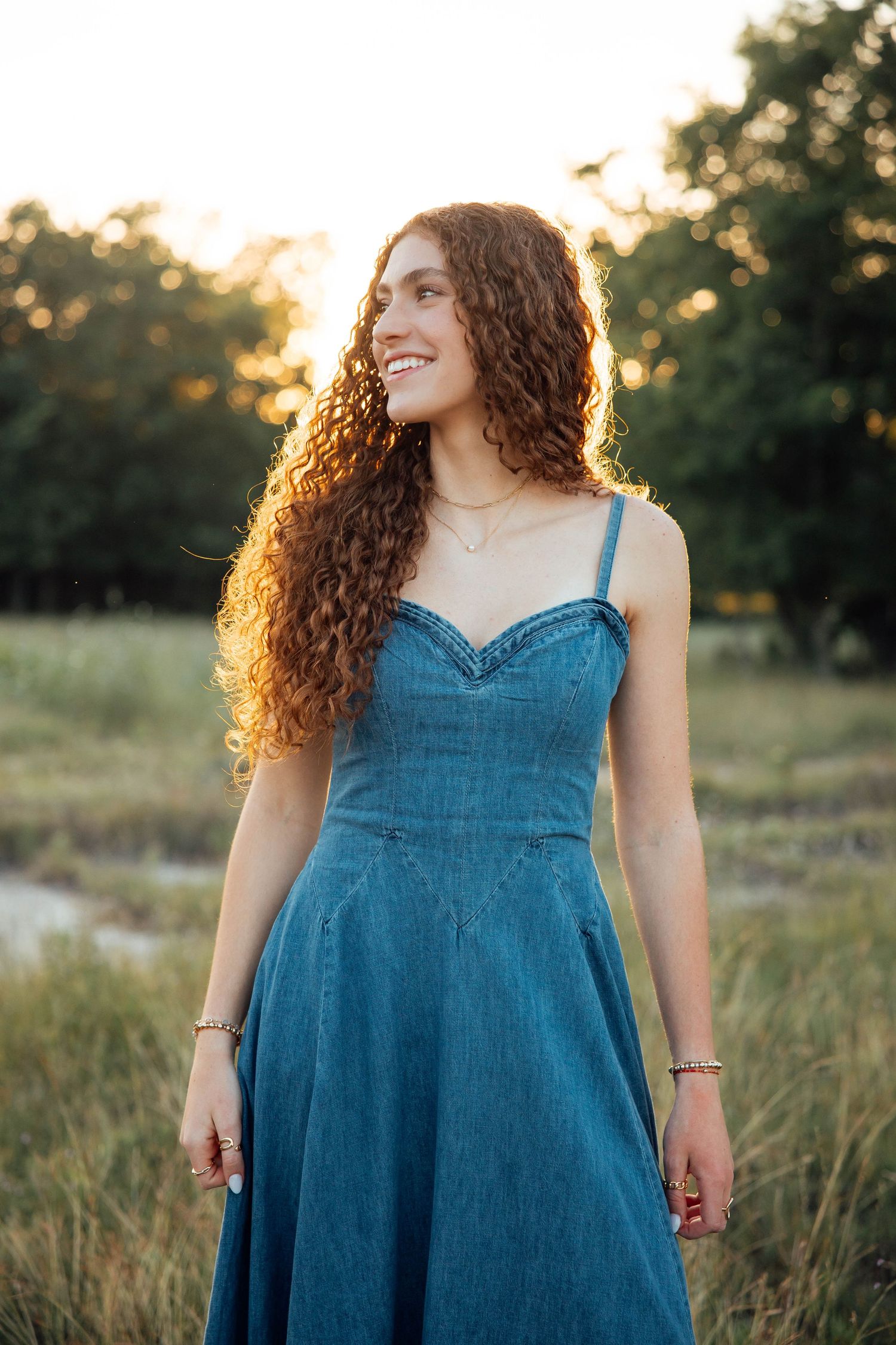 Smiling young woman with long curly red hair wearing a blue denim dress in a sunlit field.