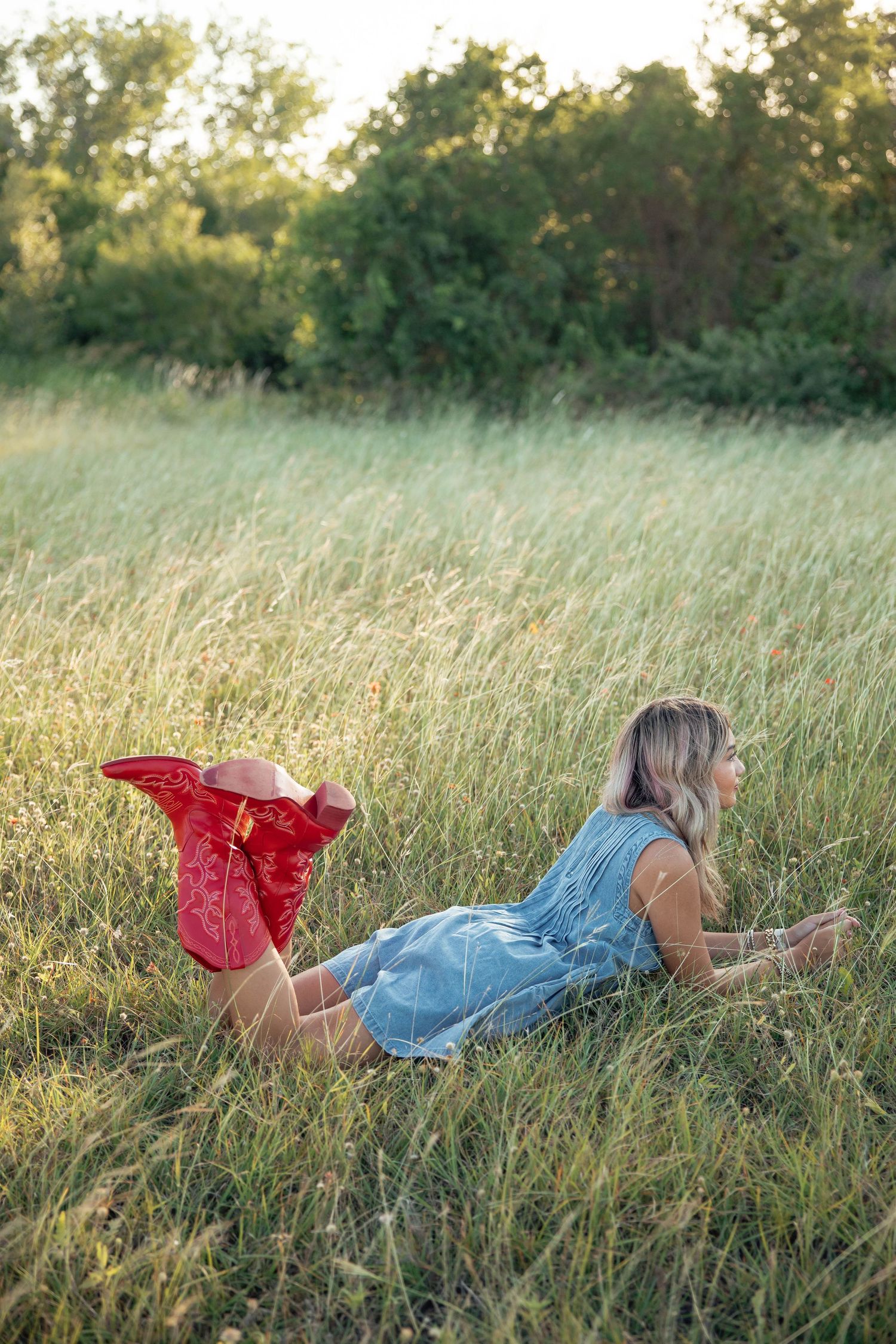 Blonde woman in blue denim dress lying in tall green grass field wearing red cowboy boots.