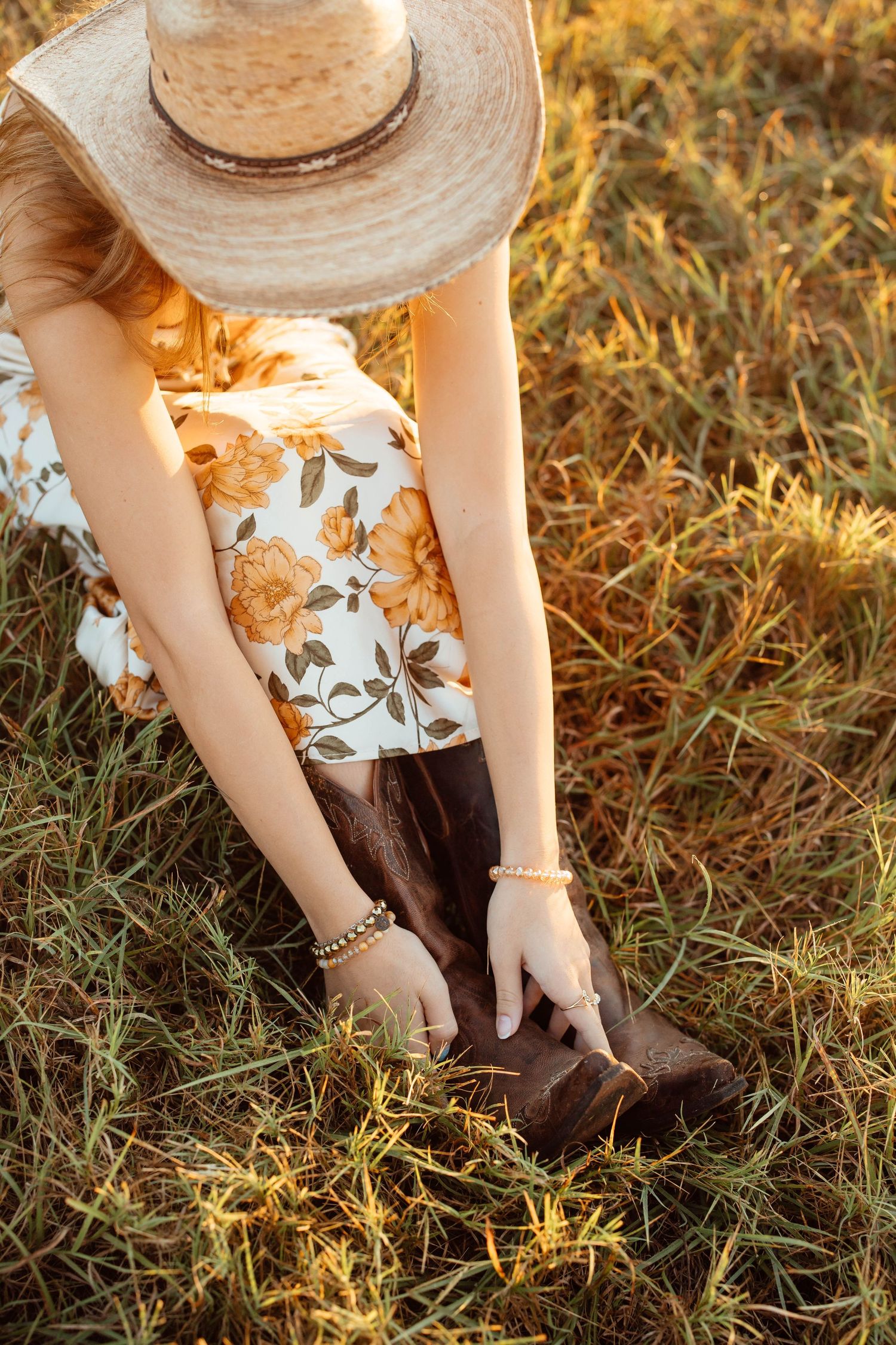 A woman in a floral dress and straw hat adjusts brown cowboy boots while sitting in a grassy field.