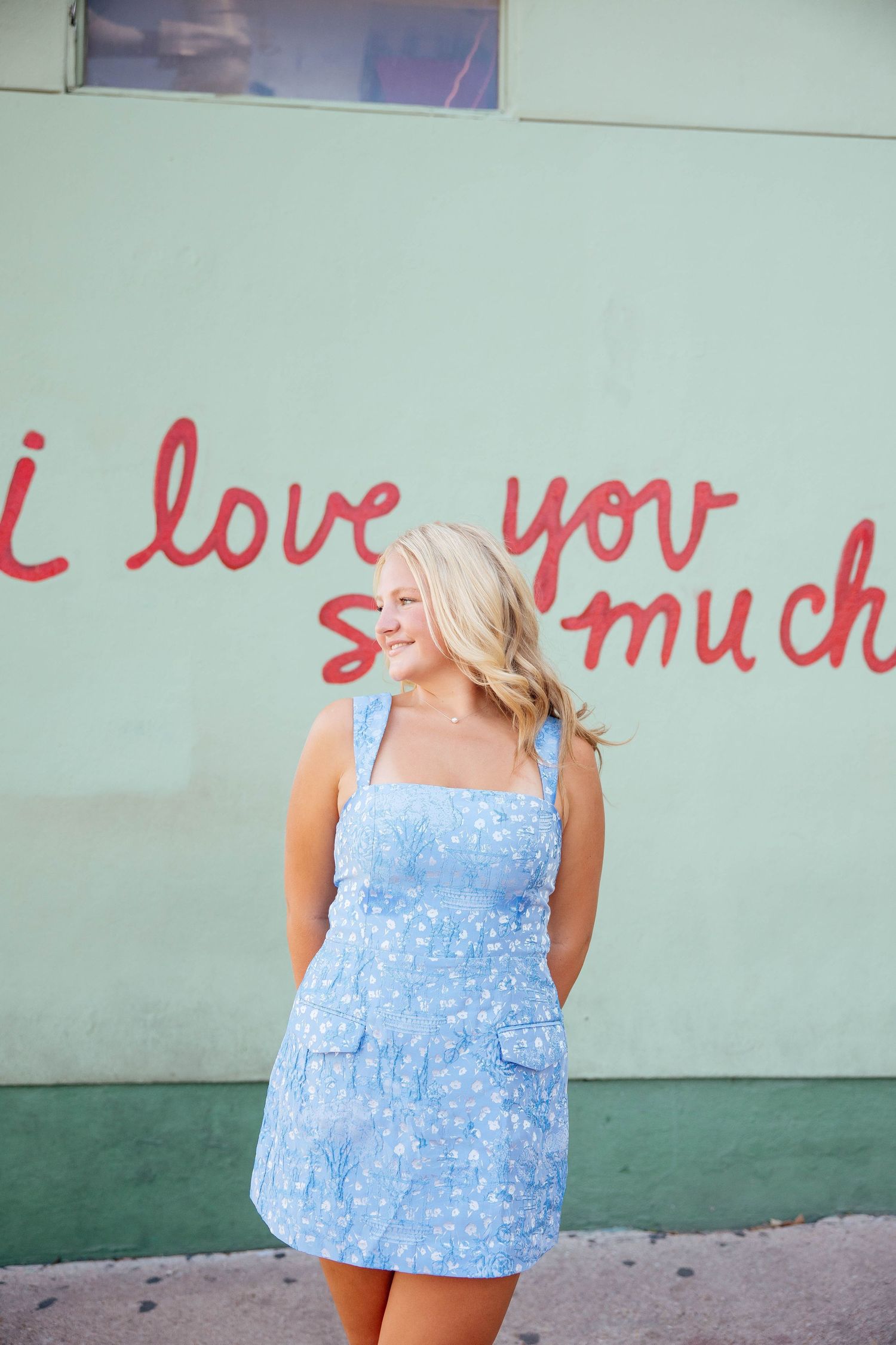 Blonde woman in blue floral sundress standing before green wall with red love quote mural.