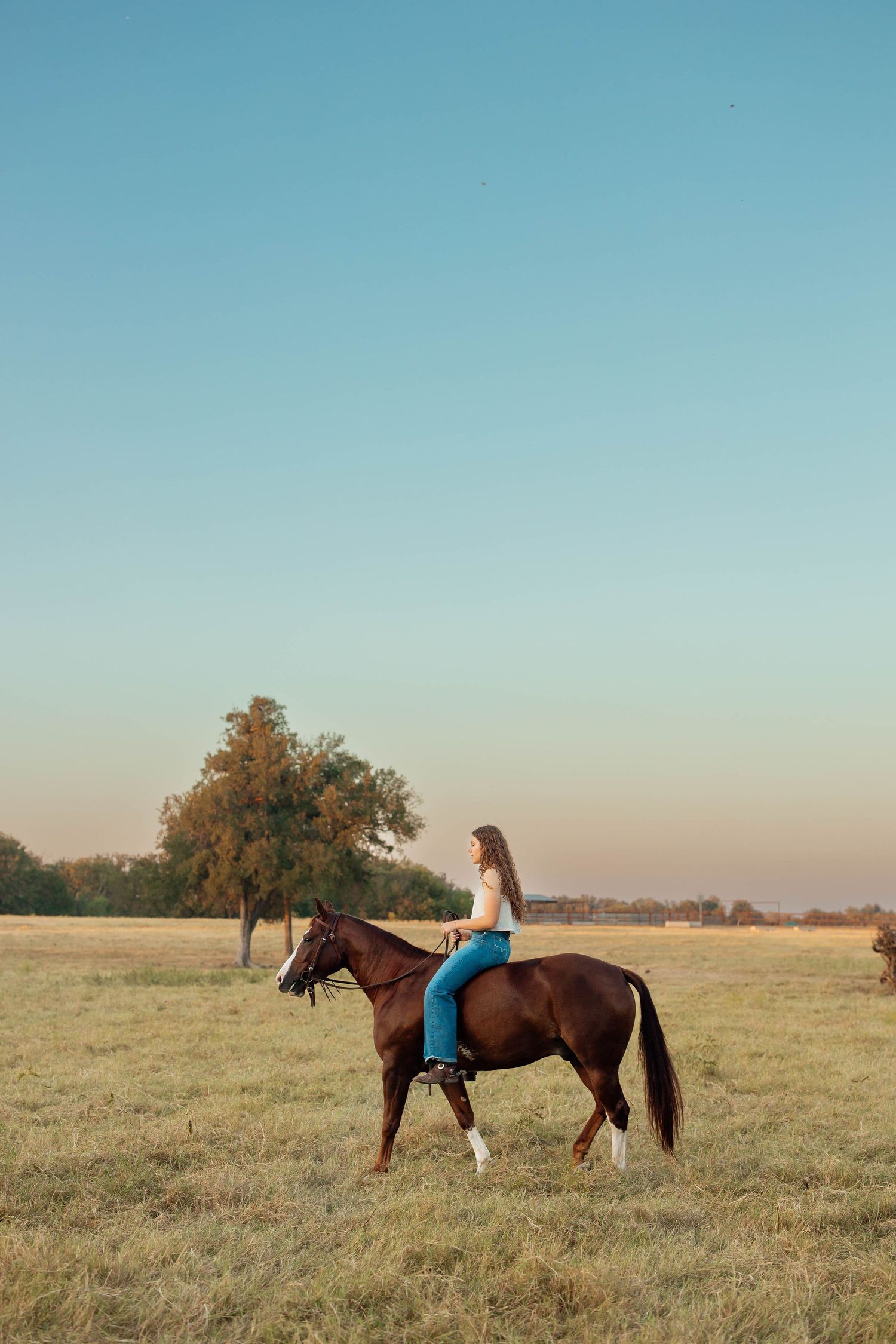 Young woman in white top and blue jeans riding a brown horse across an open field at dusk.