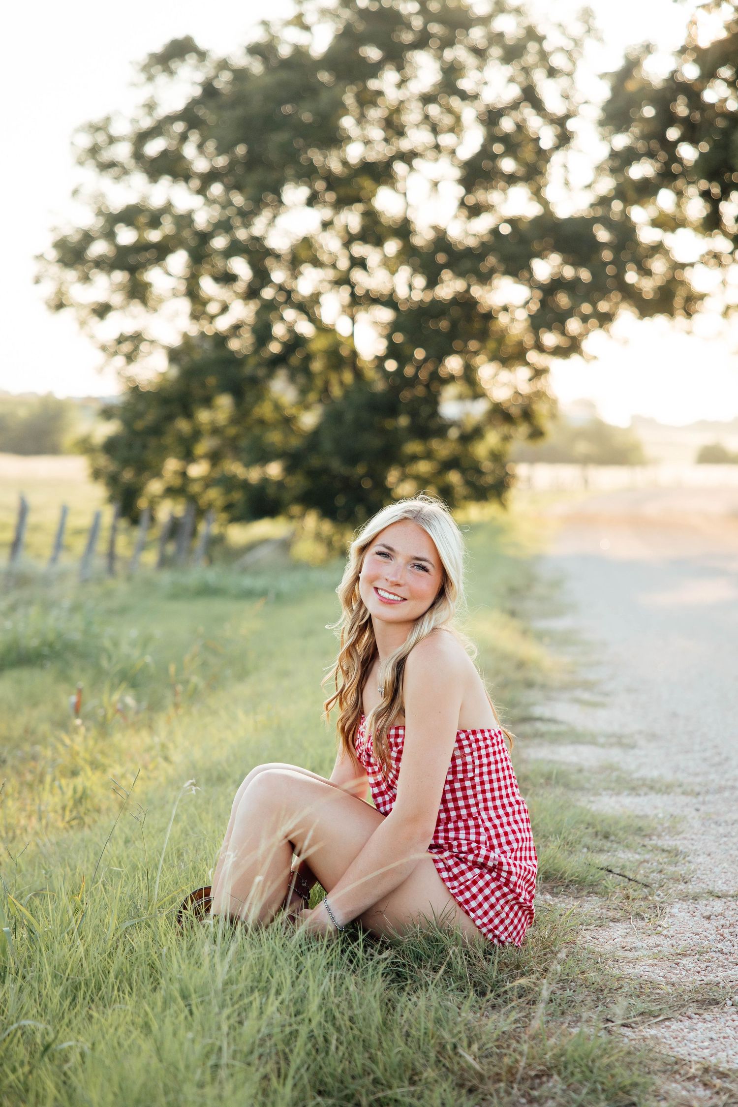 Smiling blonde girl in red gingham romper sits relaxed on grassy edge of a sunlit country road.