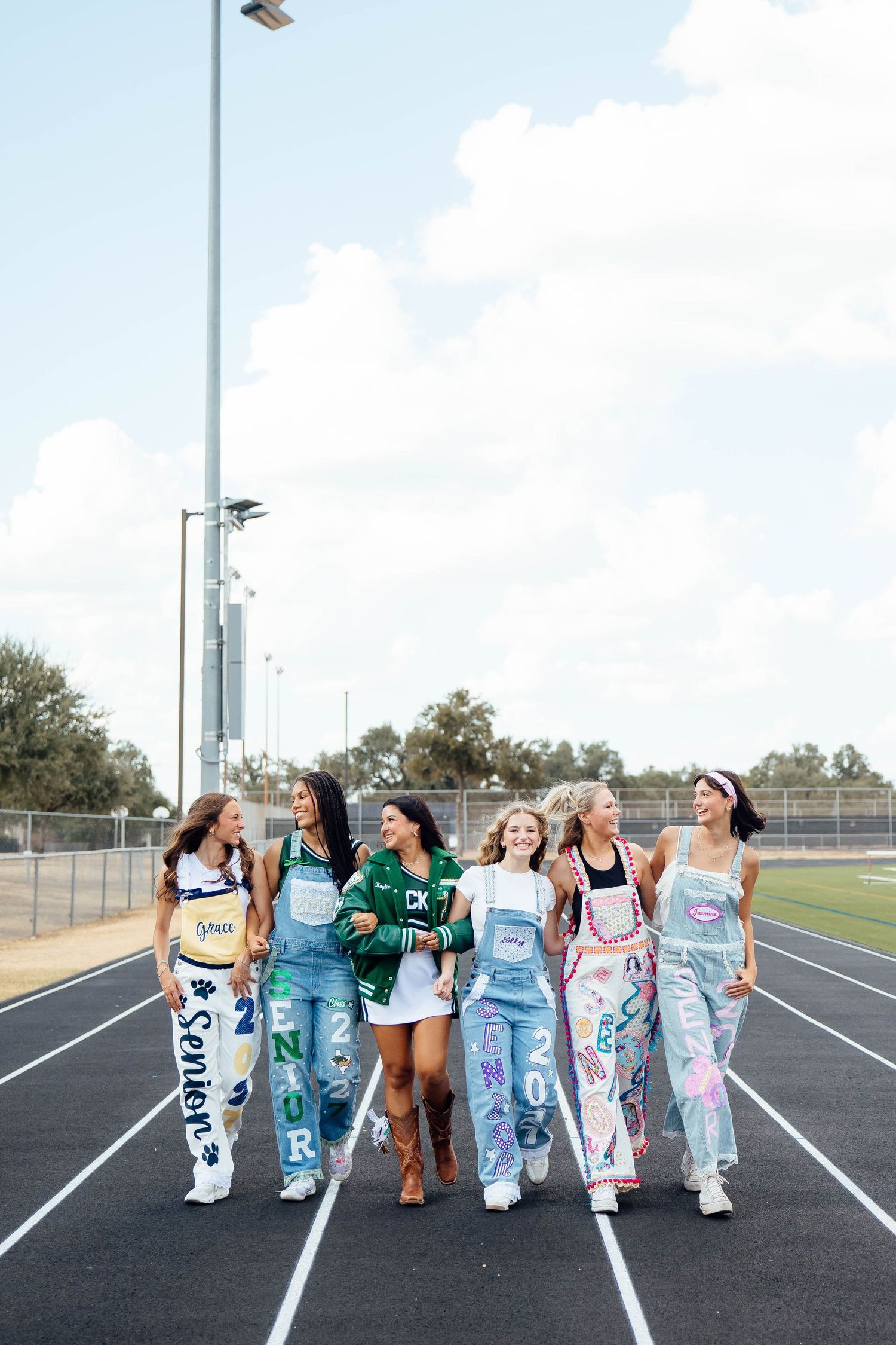 Six teen girls in painted senior overalls walk together on a school track under blue sky.
