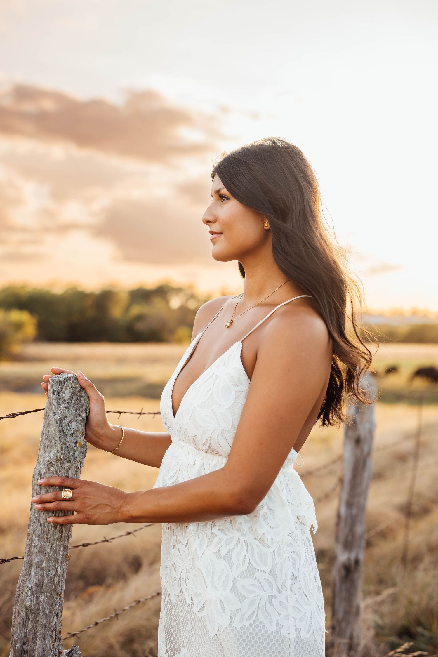Young woman in white lace dress standing by a rustic fence post at golden hour sunset.