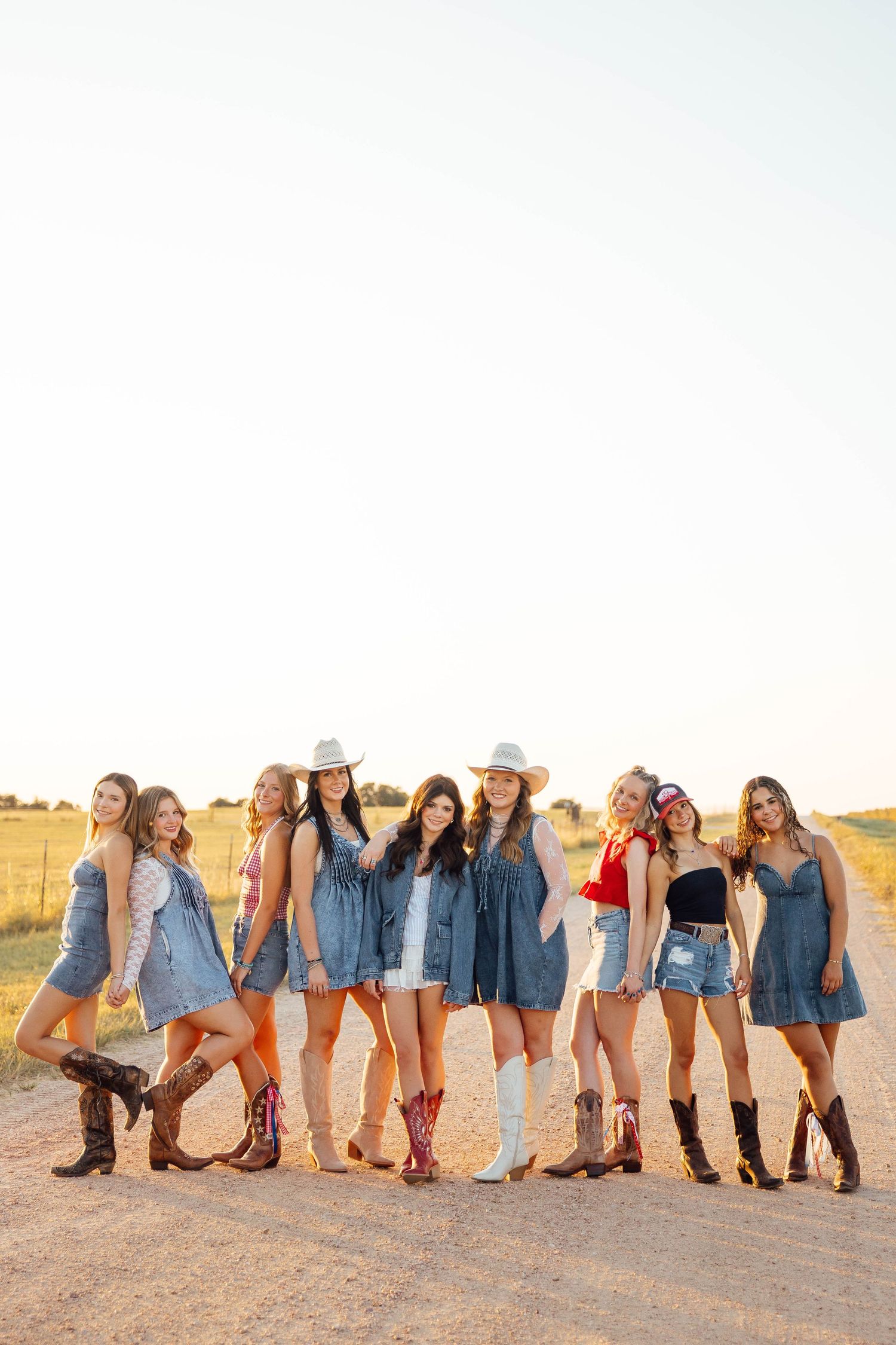 Group of young women in denim outfits and cowboy boots posing on a dusty dirt road at sunset.