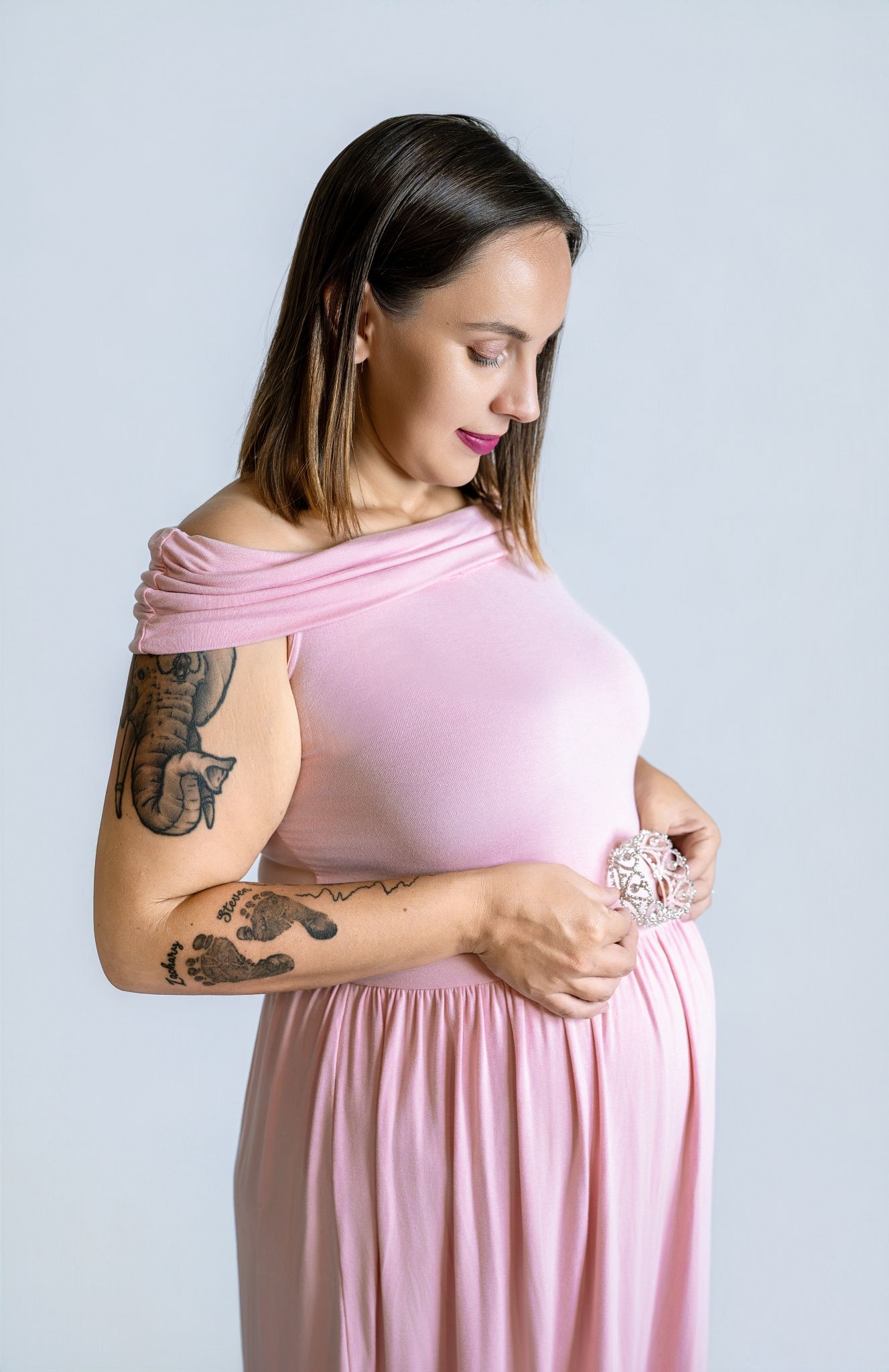 Pregnant woman in an off-shoulder pink dress with arm tattoos gently cradles her baby bump while looking down against a light backdrop.