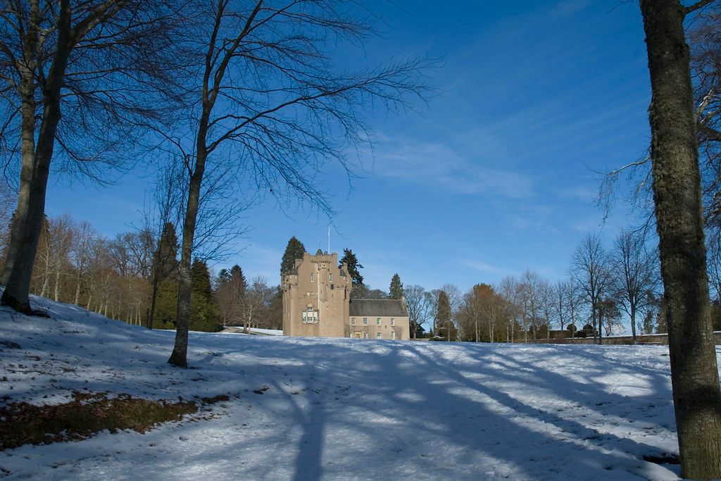Historic stone castle surrounded by snow-covered grounds and bare winter trees under a bright blue sky.