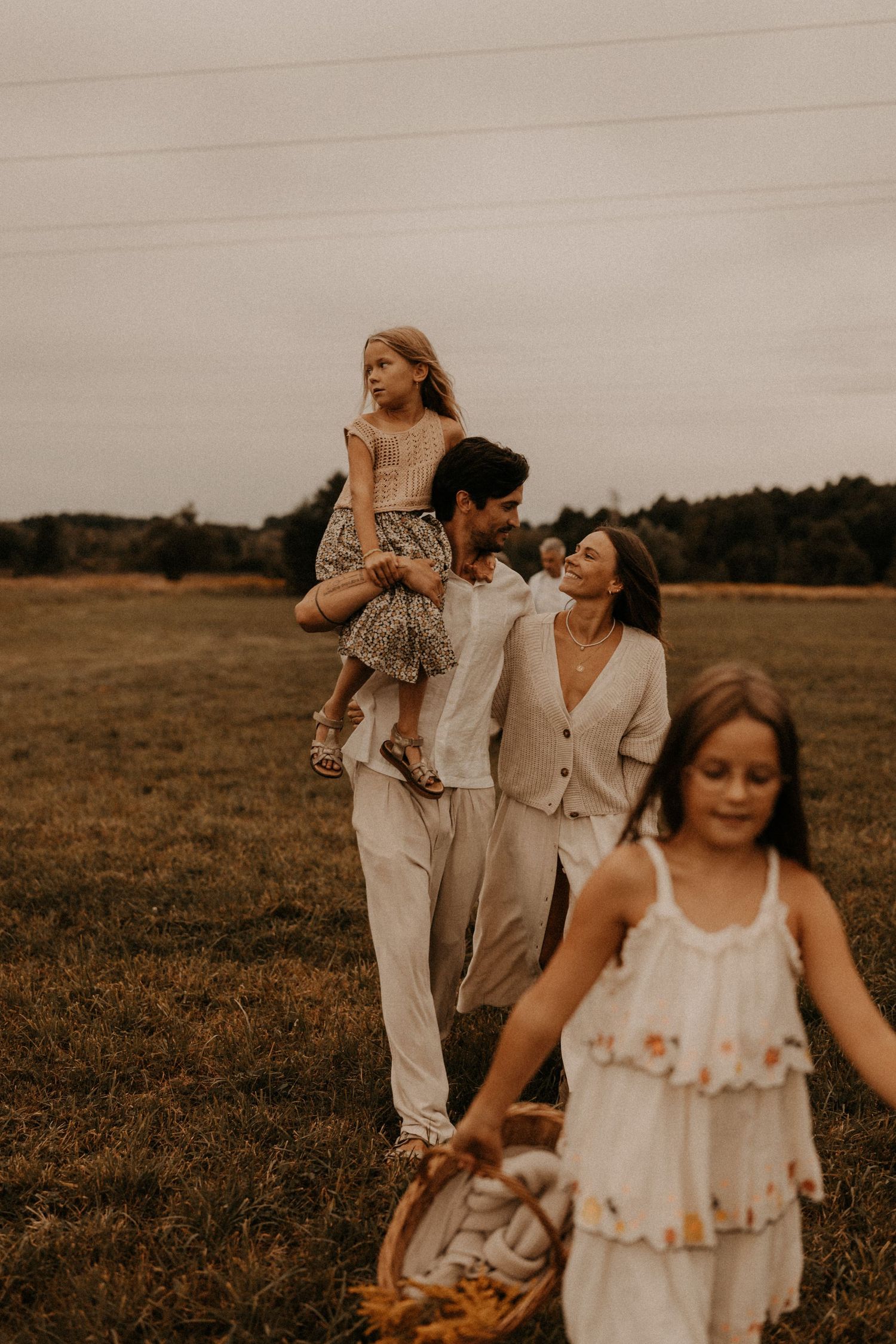 Family walking in a field, parents carrying daughter on shoulders while another girl leads with a basket.