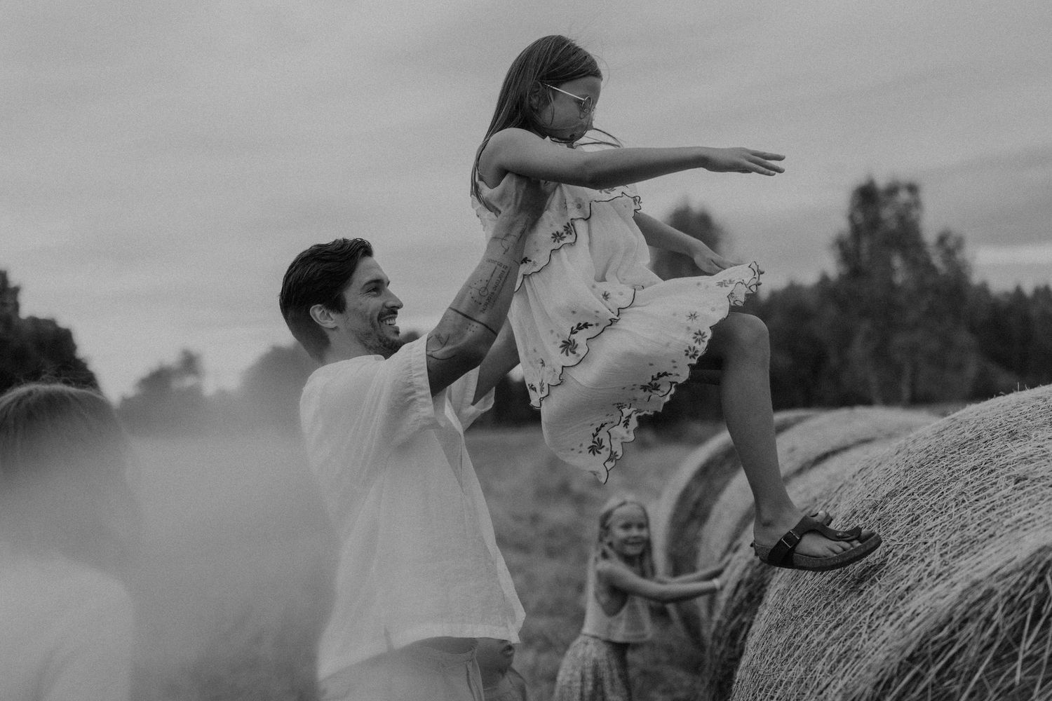 Black and white photo of a man lifting a young girl on a hay bale in a rural field setting.