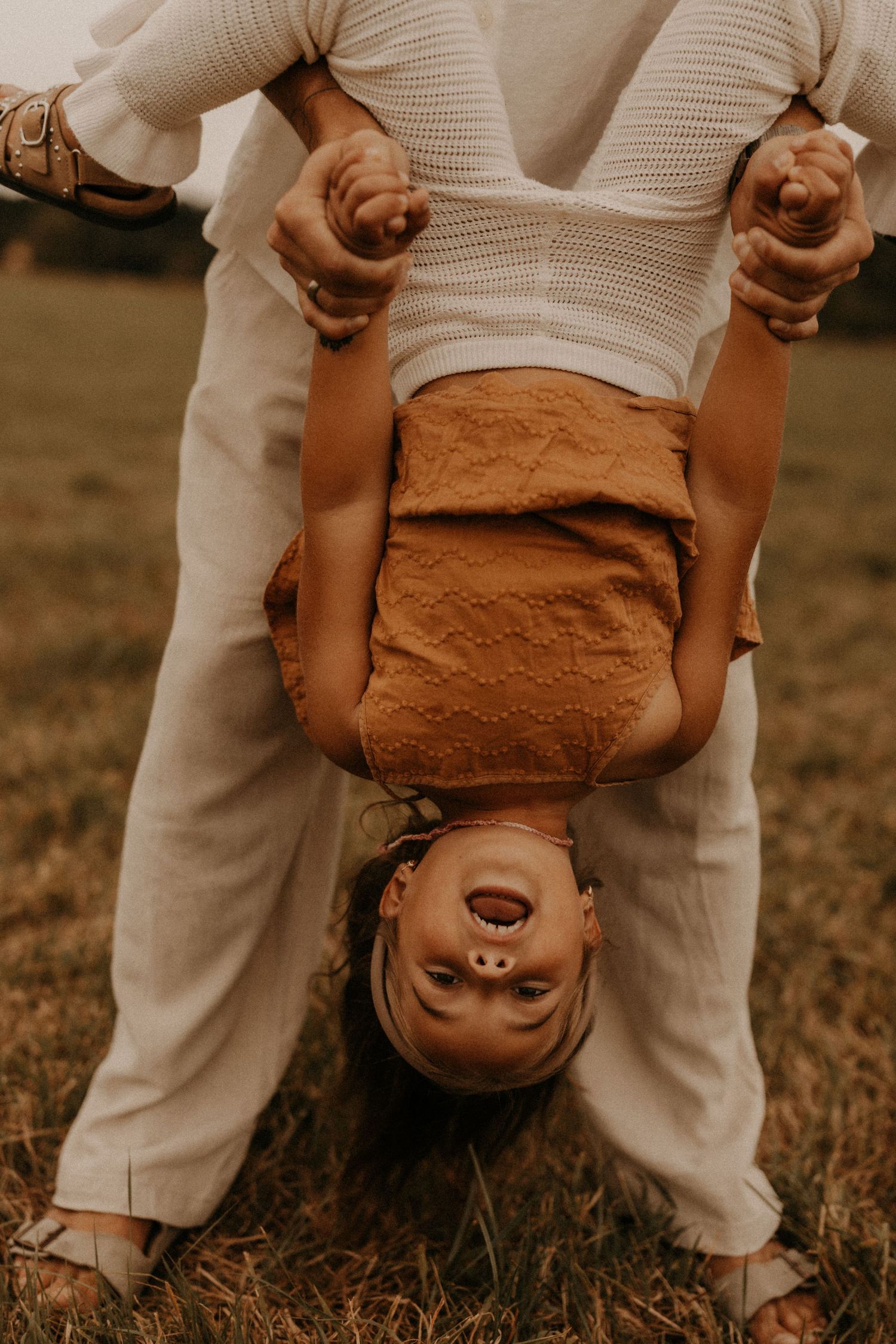 Adult playfully holds child upside down by hands in grassy field, both wearing warm neutral tones outdoors.