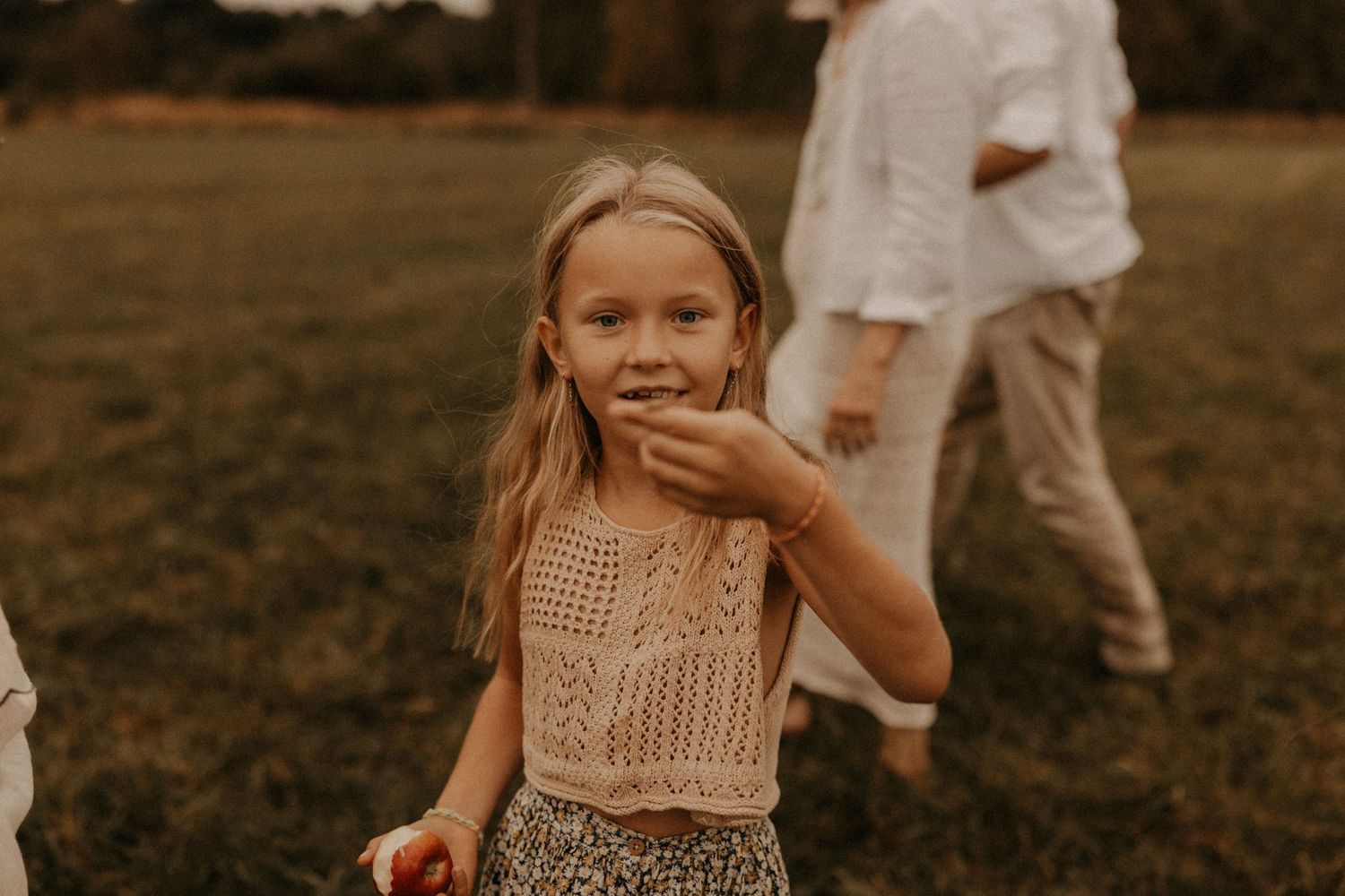 Young blonde girl in floral skirt and pink top smiling outdoors in a grassy field during golden hour.