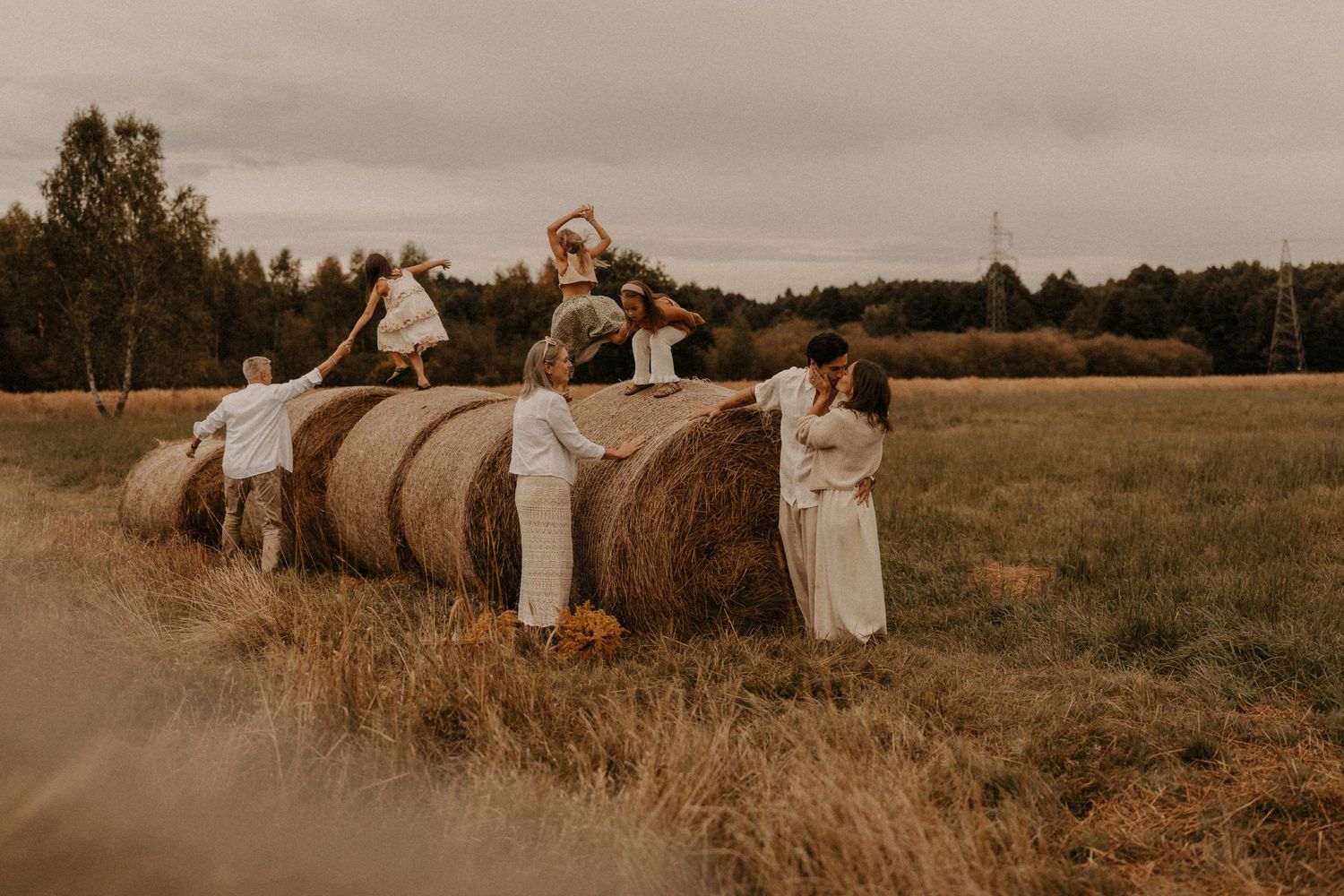Family in white clothing playing around a large hay bale in an open field during golden hour.