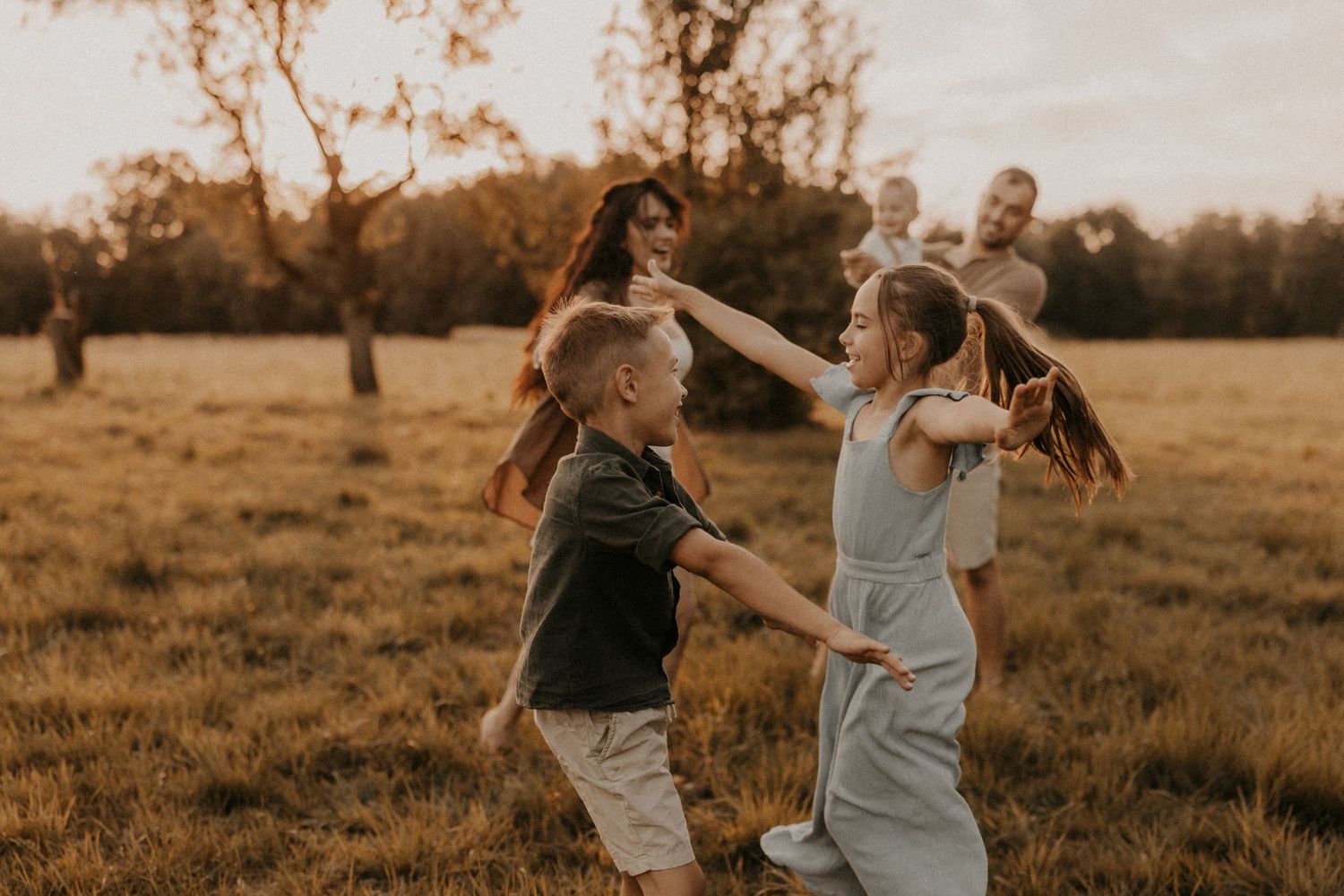 Two children play and spin together in a sunlit field while their parents watch in the background.sesje zdjęciowe Rybnik, Wodzisław Śląski