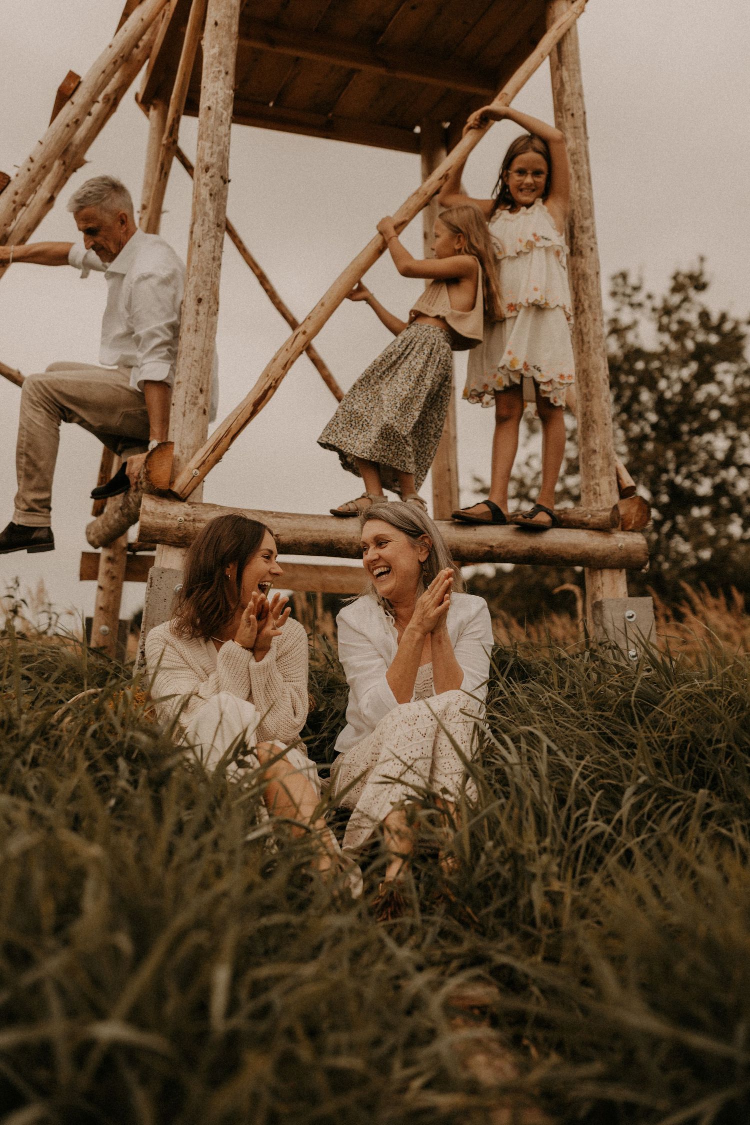 Multi-generational family enjoying outdoor time near a rustic wooden structure in a green meadow.