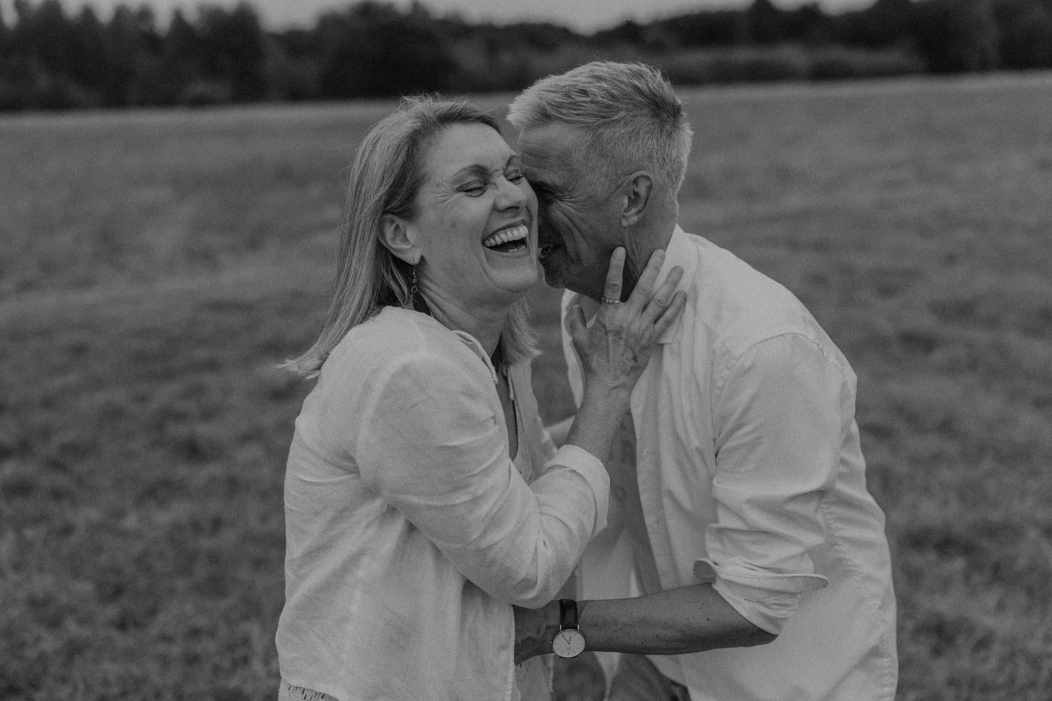 Black and white photo of a laughing couple embracing in a field, sharing a joyful intimate moment outdoors.