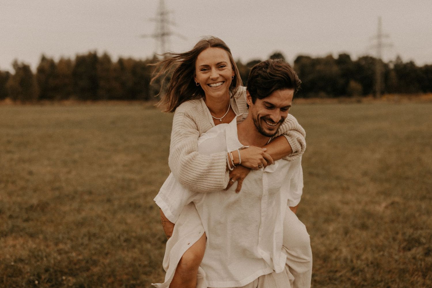 A joyful couple in white outfits shares a piggyback ride in a green field with trees and power lines behind them.