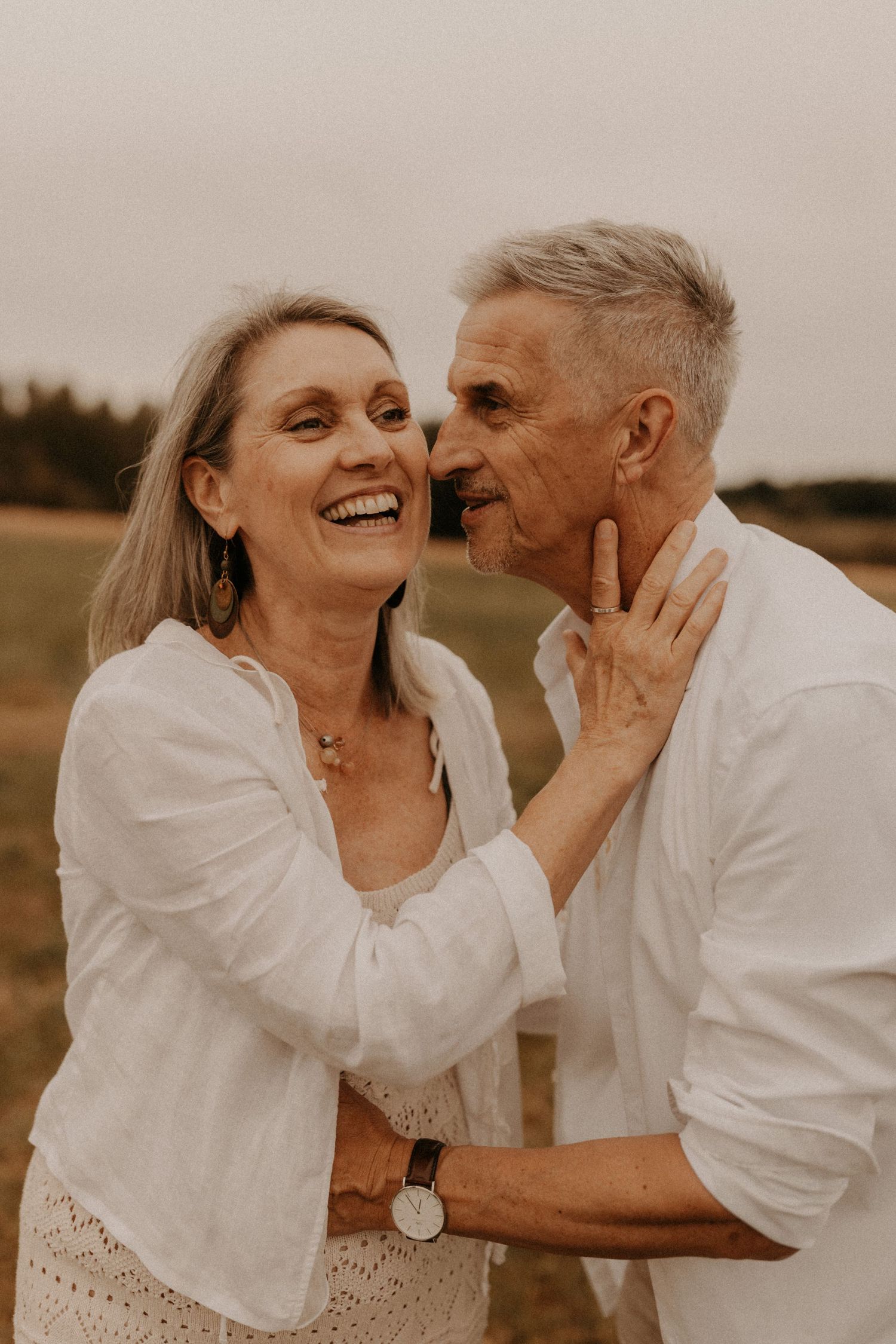 A joyful mature couple in white linen outfits share a tender moment outdoors, laughing and embracing in a field.