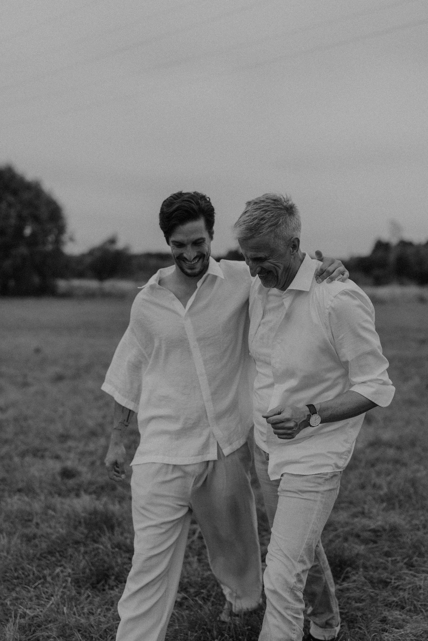 Two men in white linen outfits walk arm in arm through a field, laughing together in a black and white photo.