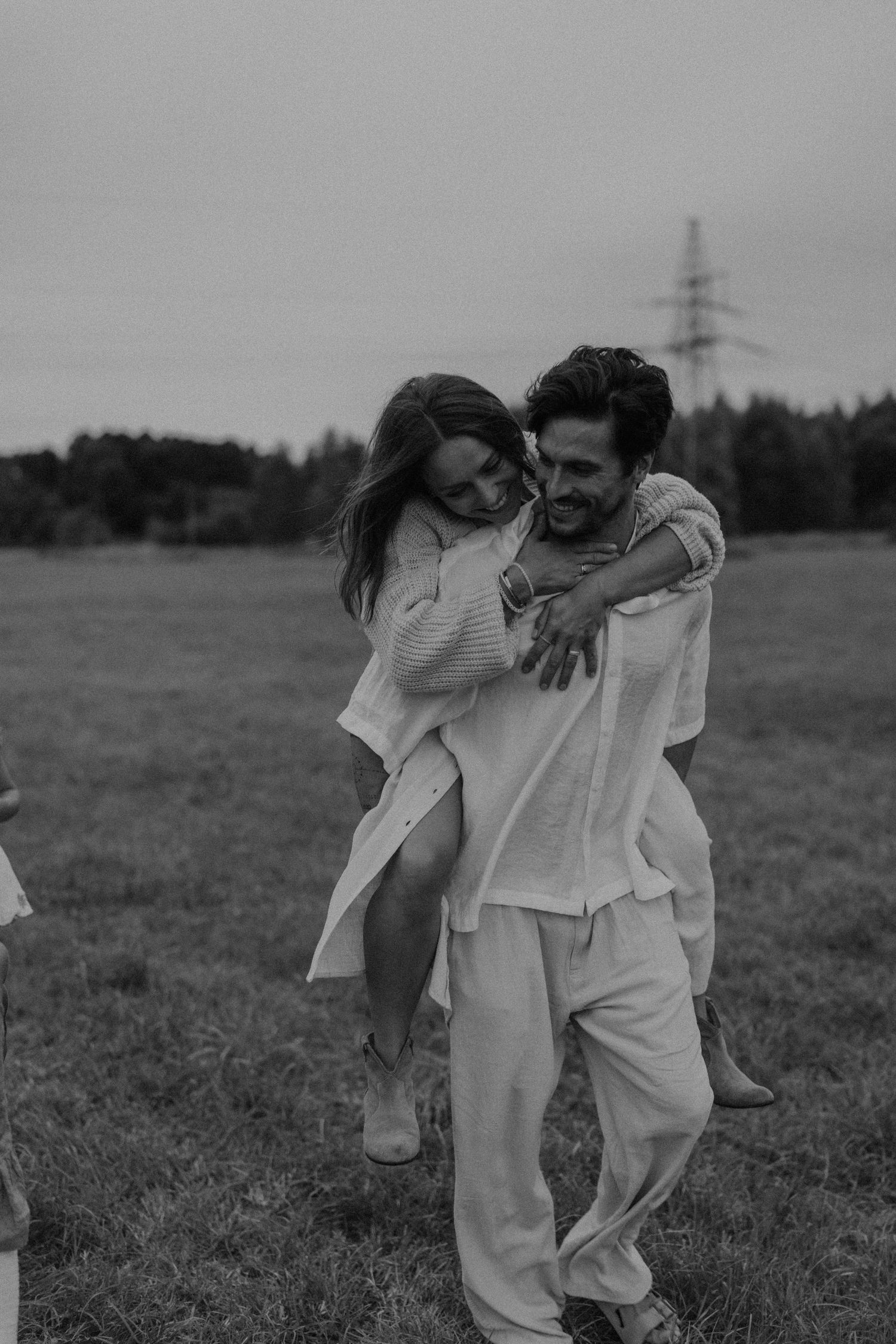 Black and white photo of a man giving a woman a piggyback ride in a field, both wearing white linen outfits.