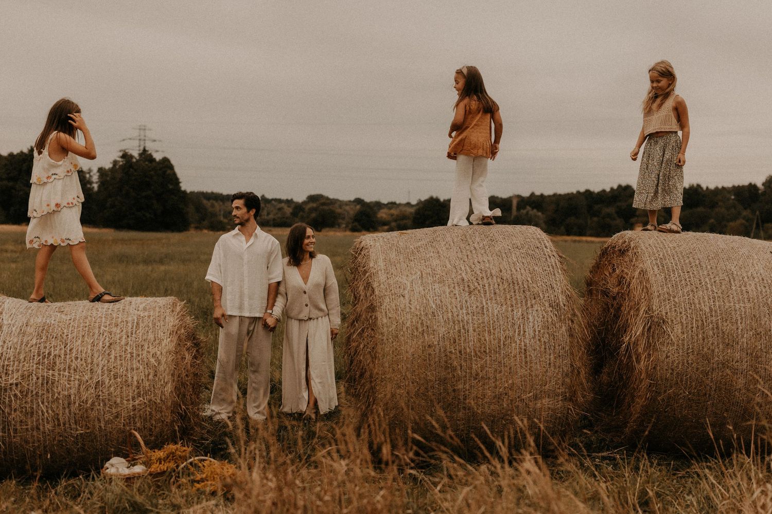 Five people pose among large hay bales in an open field, dressed in neutral tones under an overcast sky.