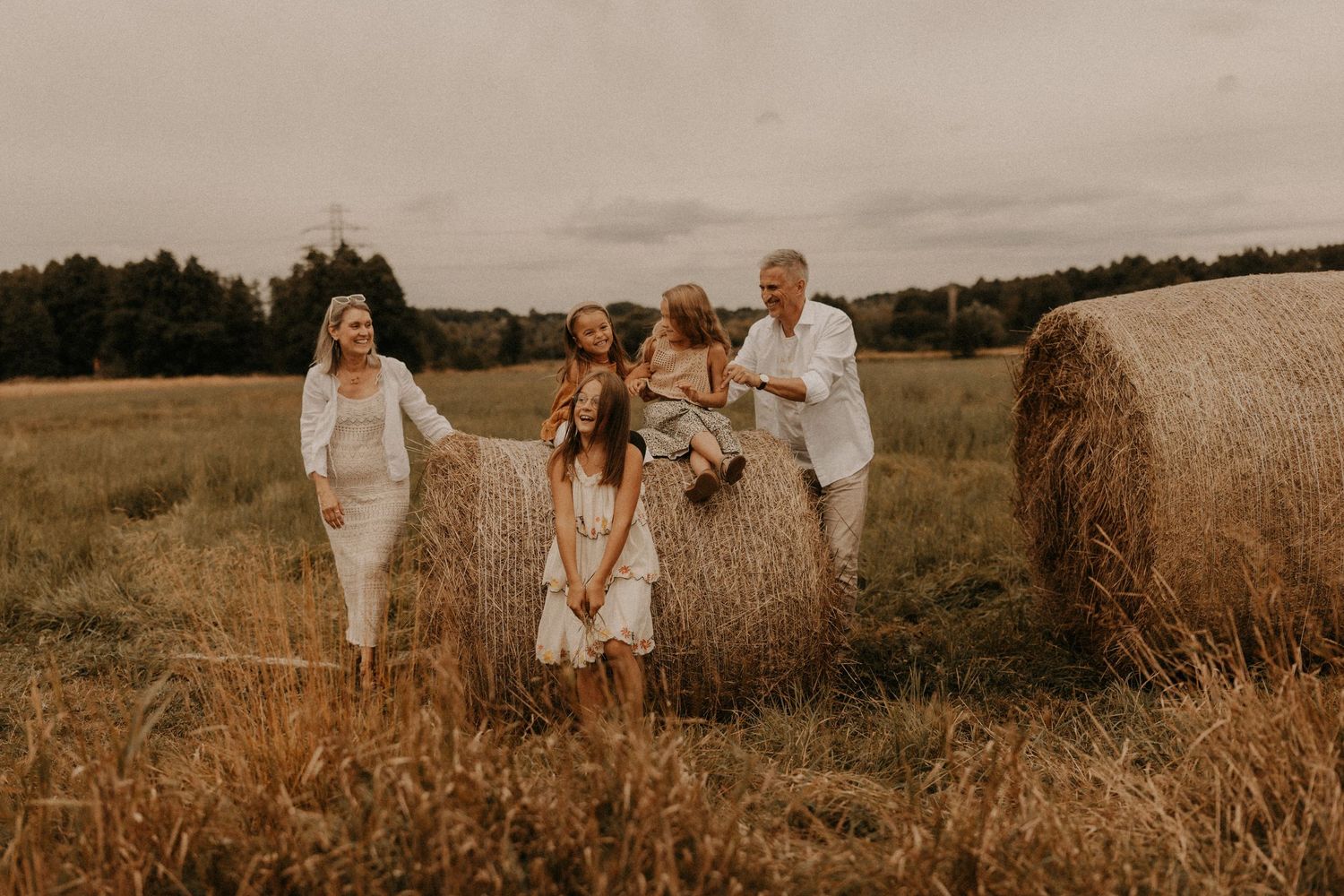 Family of four posing with hay bales in a golden field, wearing casual white and boho outfits on a cloudy day.