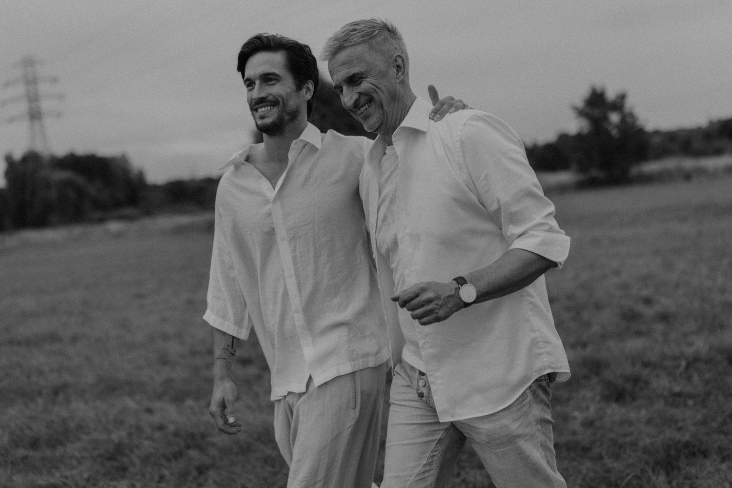 Two men in white linen shirts walk together laughing in a field, black and white photo.