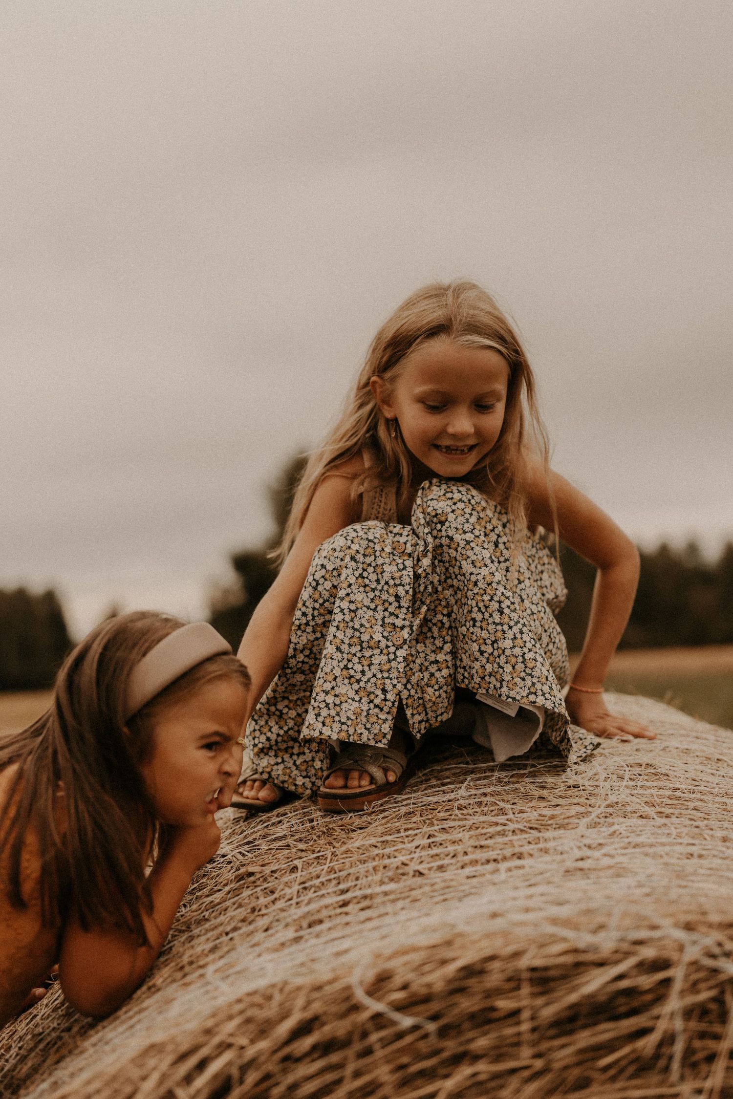 Two young girls playing on a hay bale in a field, wearing floral outfits on a cloudy day.