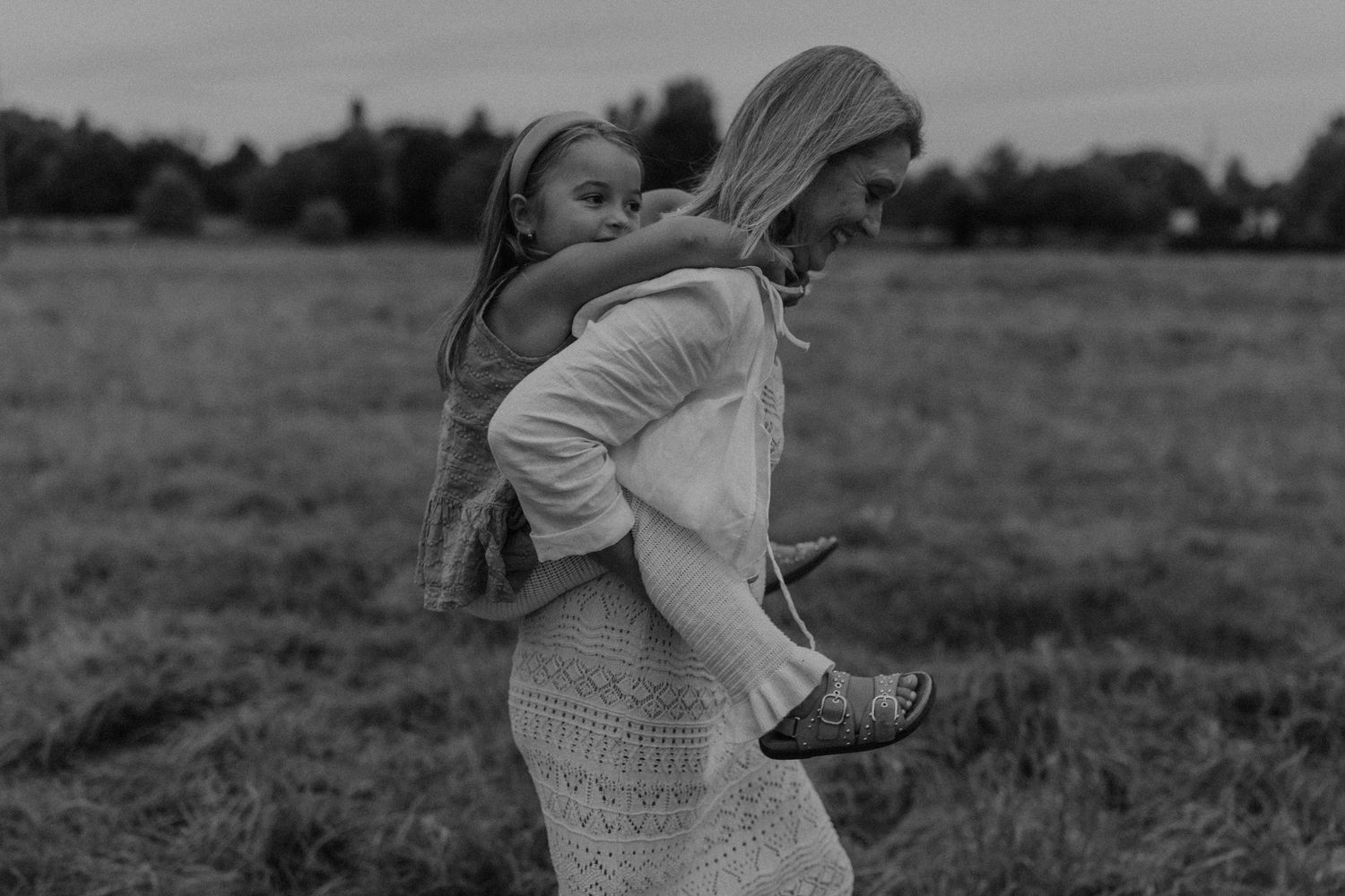 Black and white photo of a woman carrying a young girl piggyback through a grassy field.