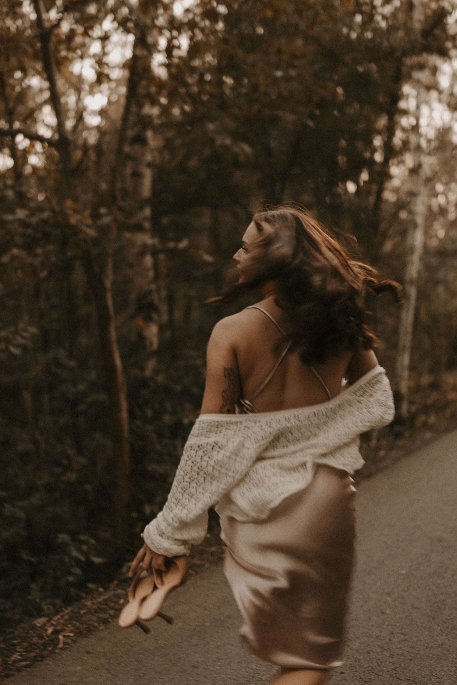 Woman in white off-shoulder sweater and satin skirt walking on forest path, hair flowing, holding heels.