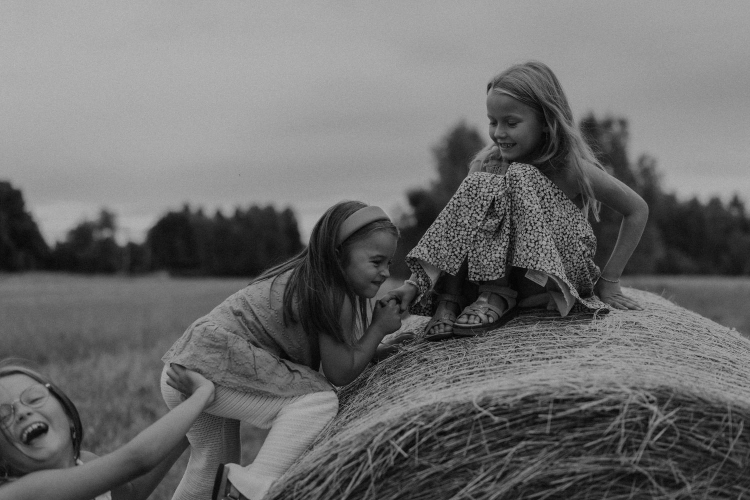 Black and white photo of three children playing on a hay bale in a field.