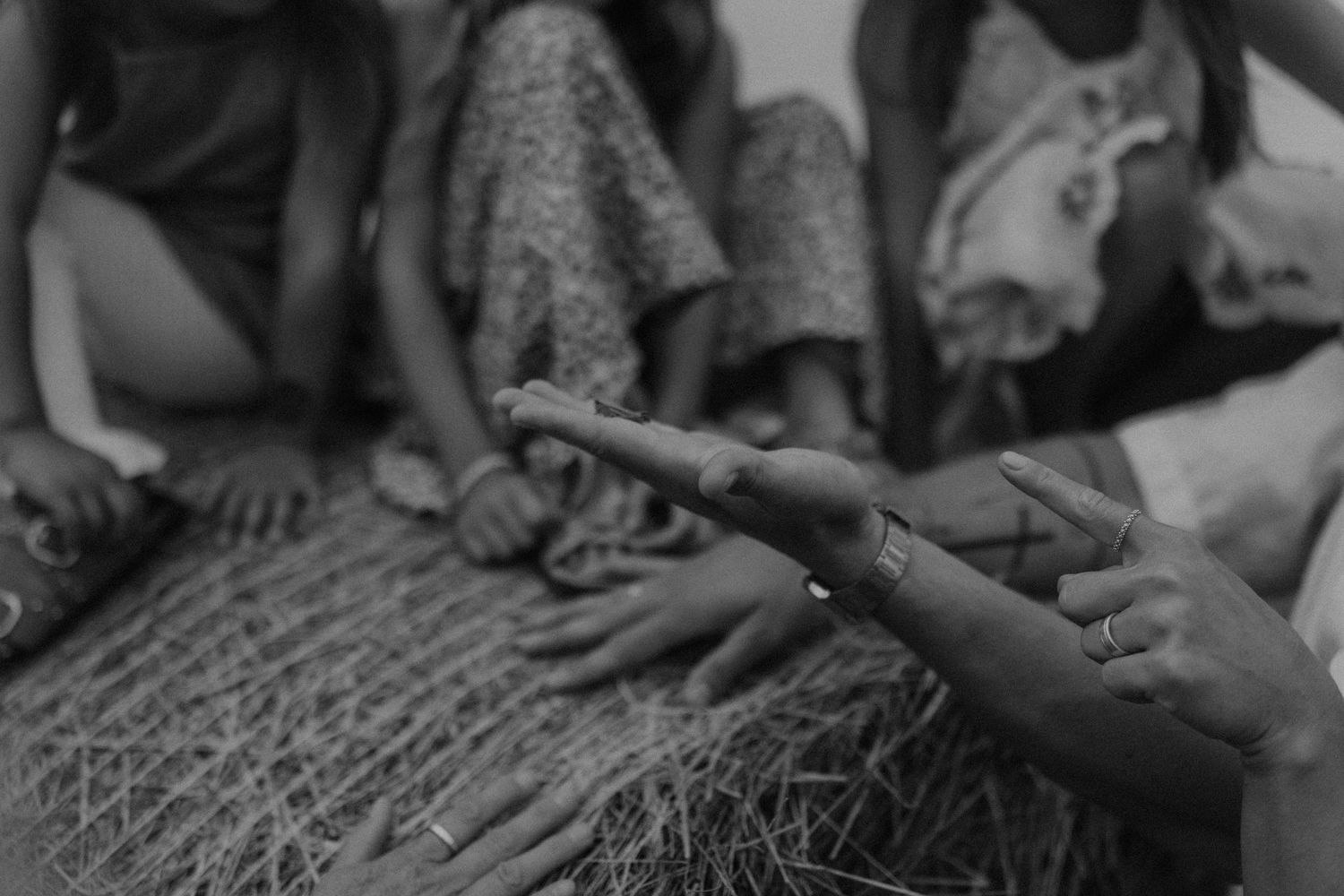 Black and white photo of children sitting together, pointing fingers at each other over woven straw mat.