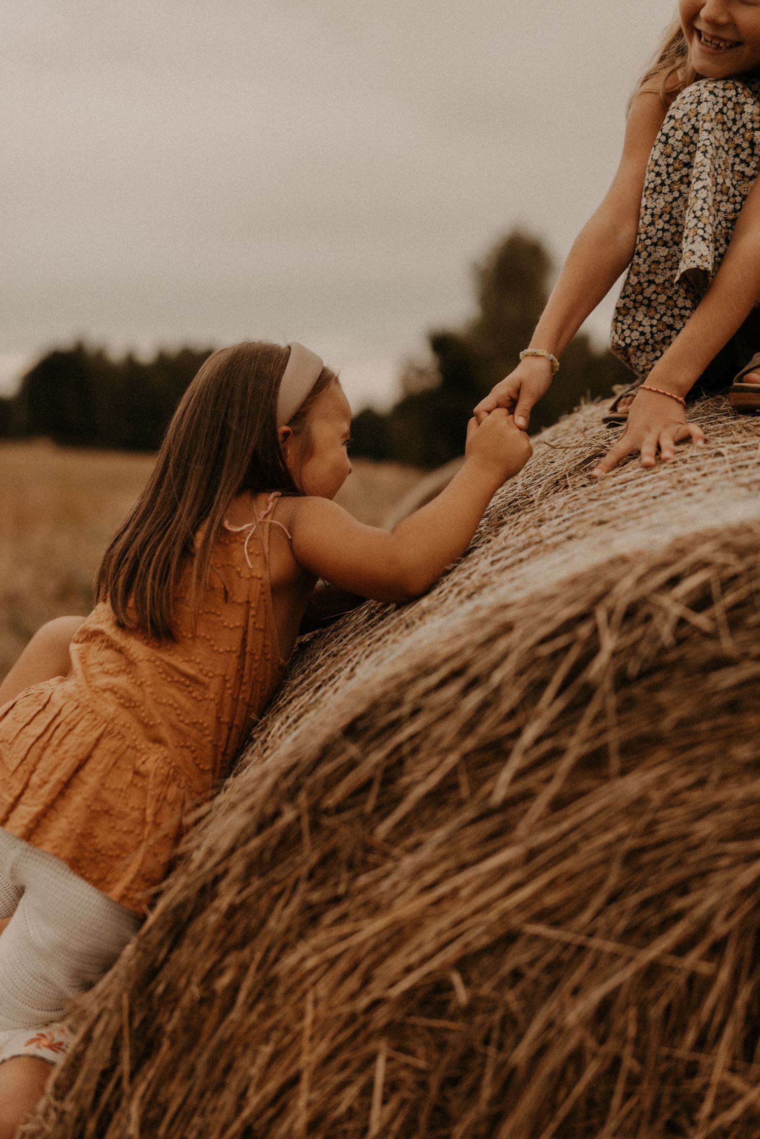 A young girl in an orange dress climbs a hay bale in a field, helped by another child on top.