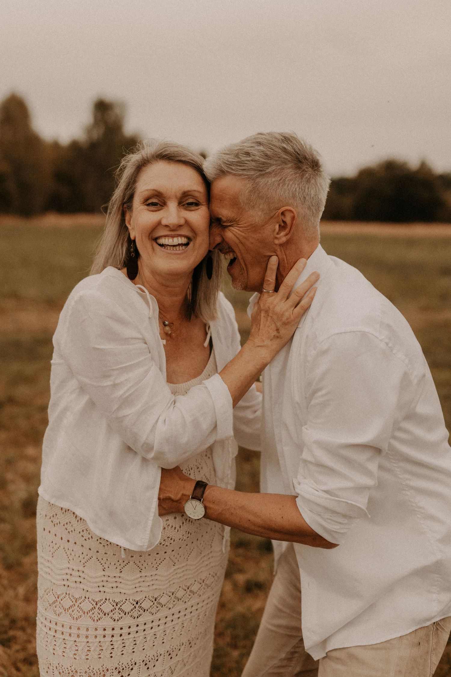 A joyful older couple in white outfits share a tender moment outdoors, as the man whispers in the laughing woman's ear.