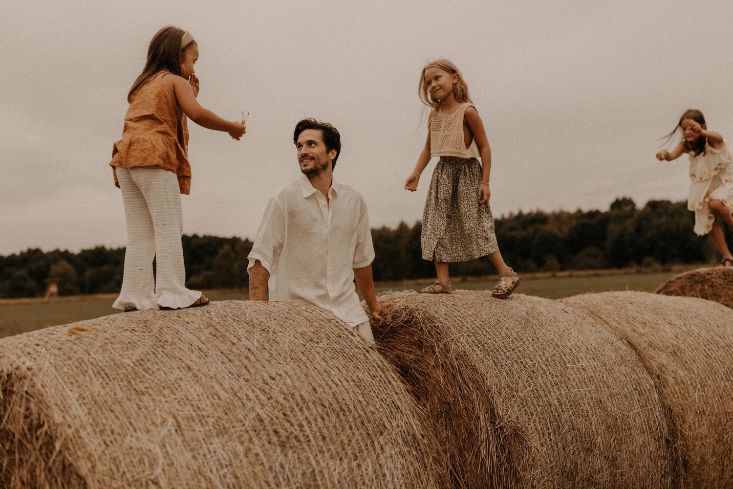 Family with children playing on large hay bales in an open field during a cloudy day.