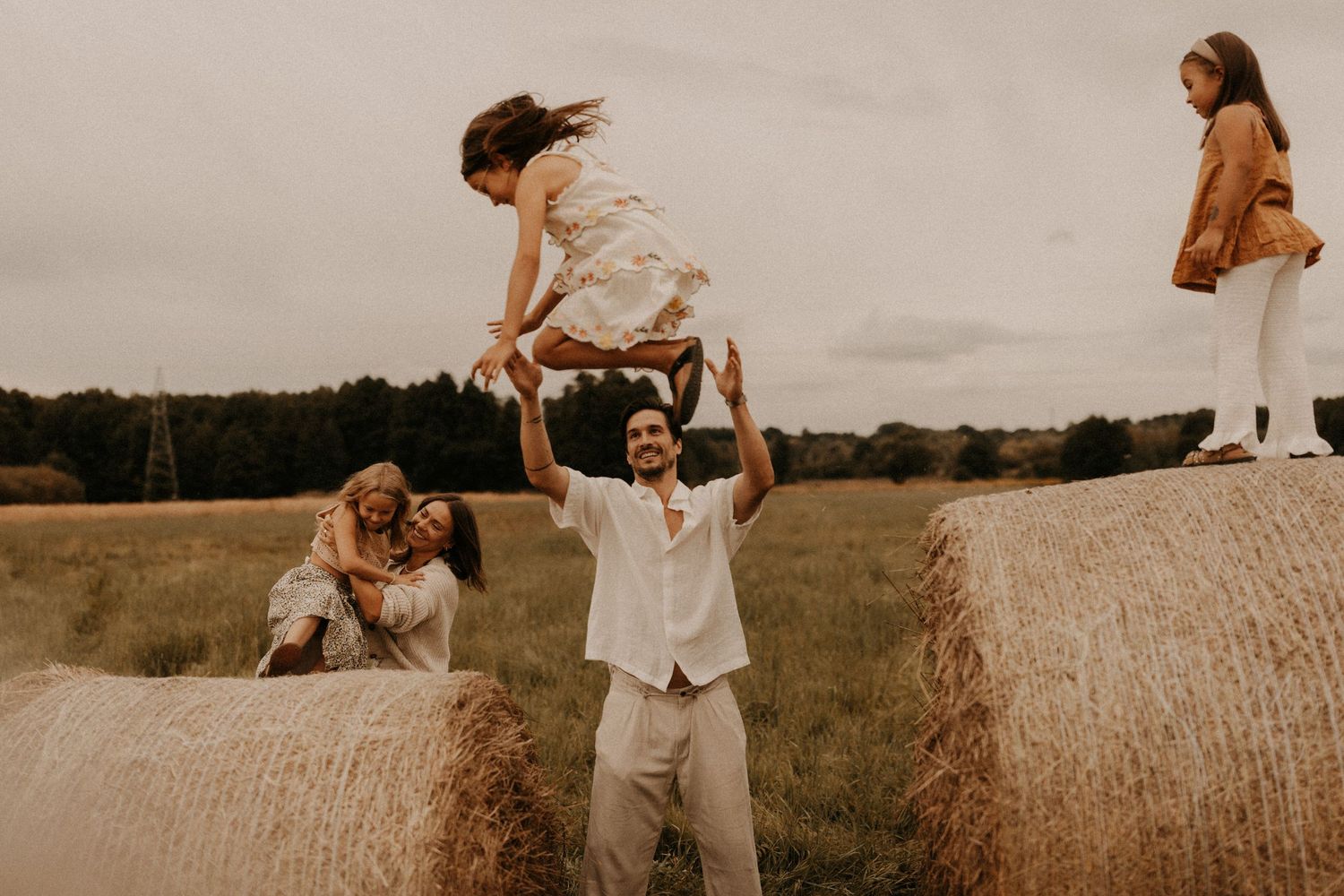 Family playing on hay bales in a field, father tossing daughter in the air, rustic outdoor setting.