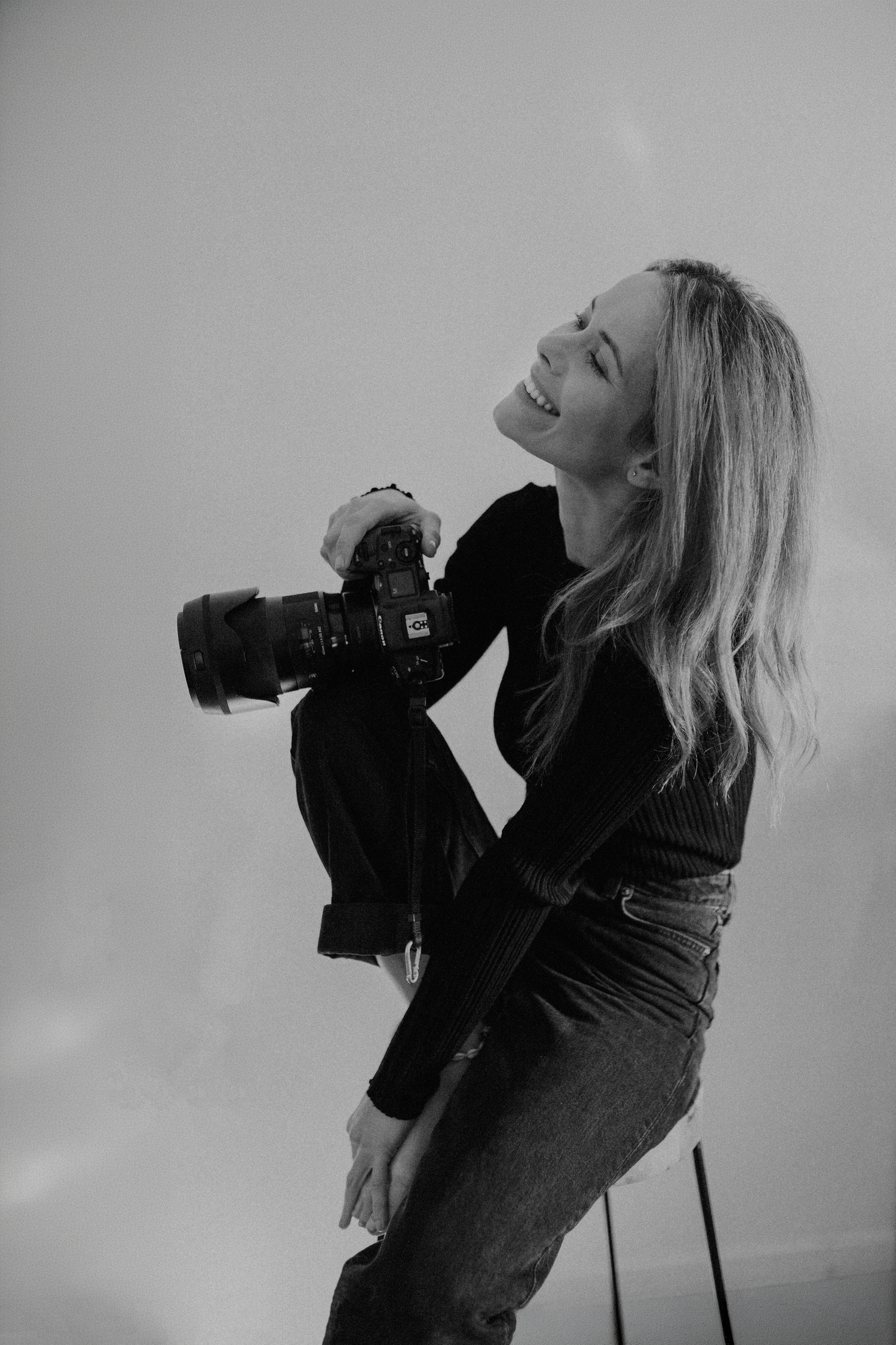 Black and white photo of a smiling blonde woman standing and holding a professional camera in a studio.