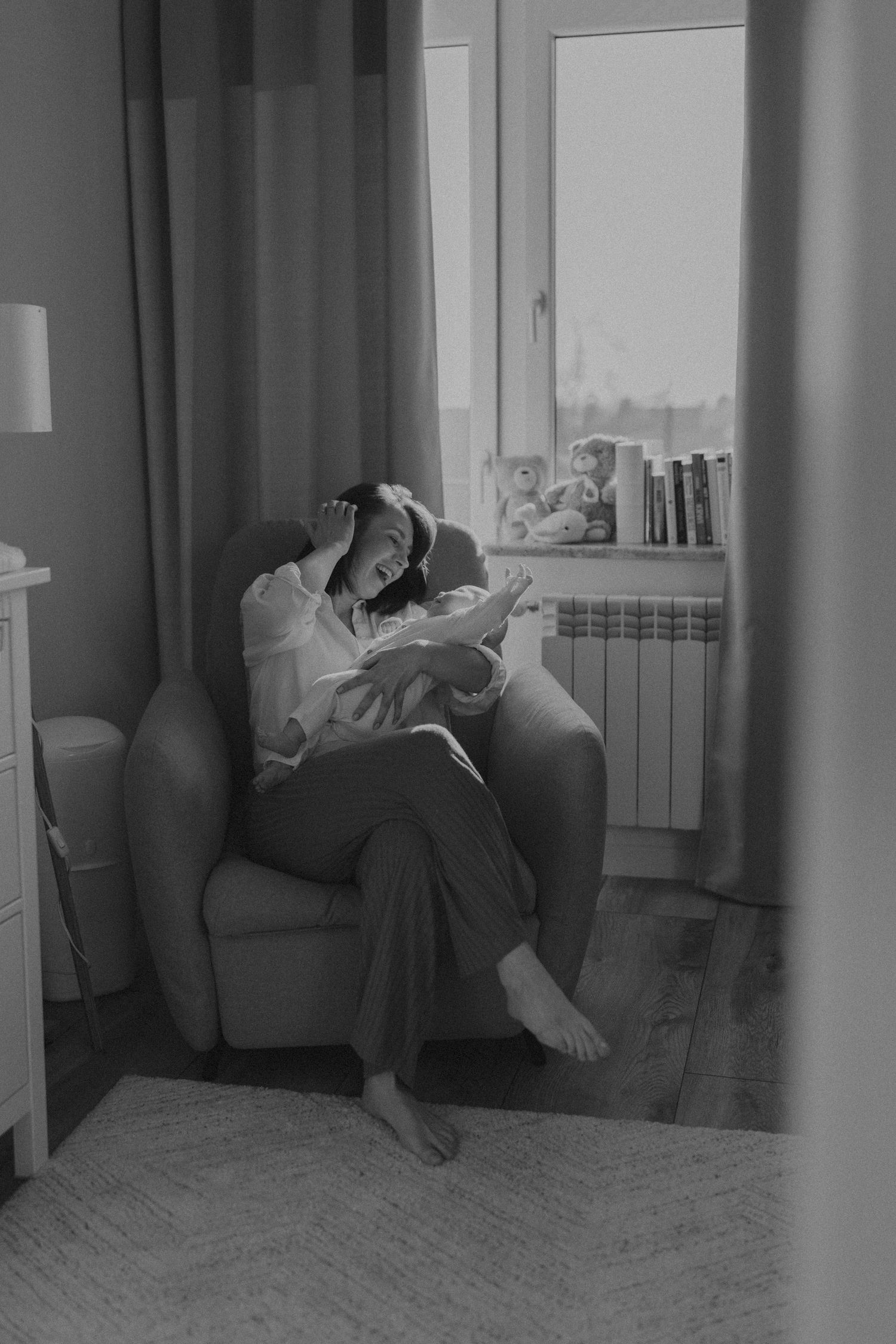 Black and white photo of a mother sitting in an armchair reading to her newborn baby by a window. sesje zdjęciowe Rybnik, Wodzisław Śląski