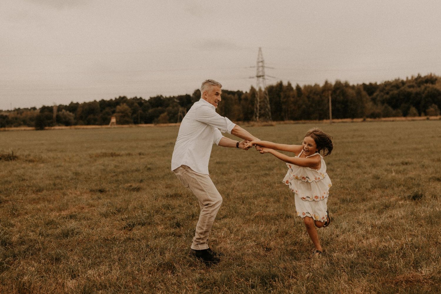 A man and young girl joyfully spin together in a vast green meadow with trees and a power line in the background.