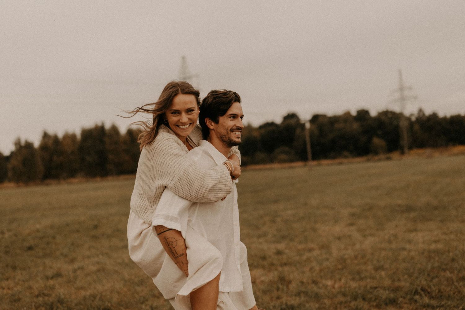 A joyful couple in white outfits shares a piggyback ride in a green meadow with trees in the background.